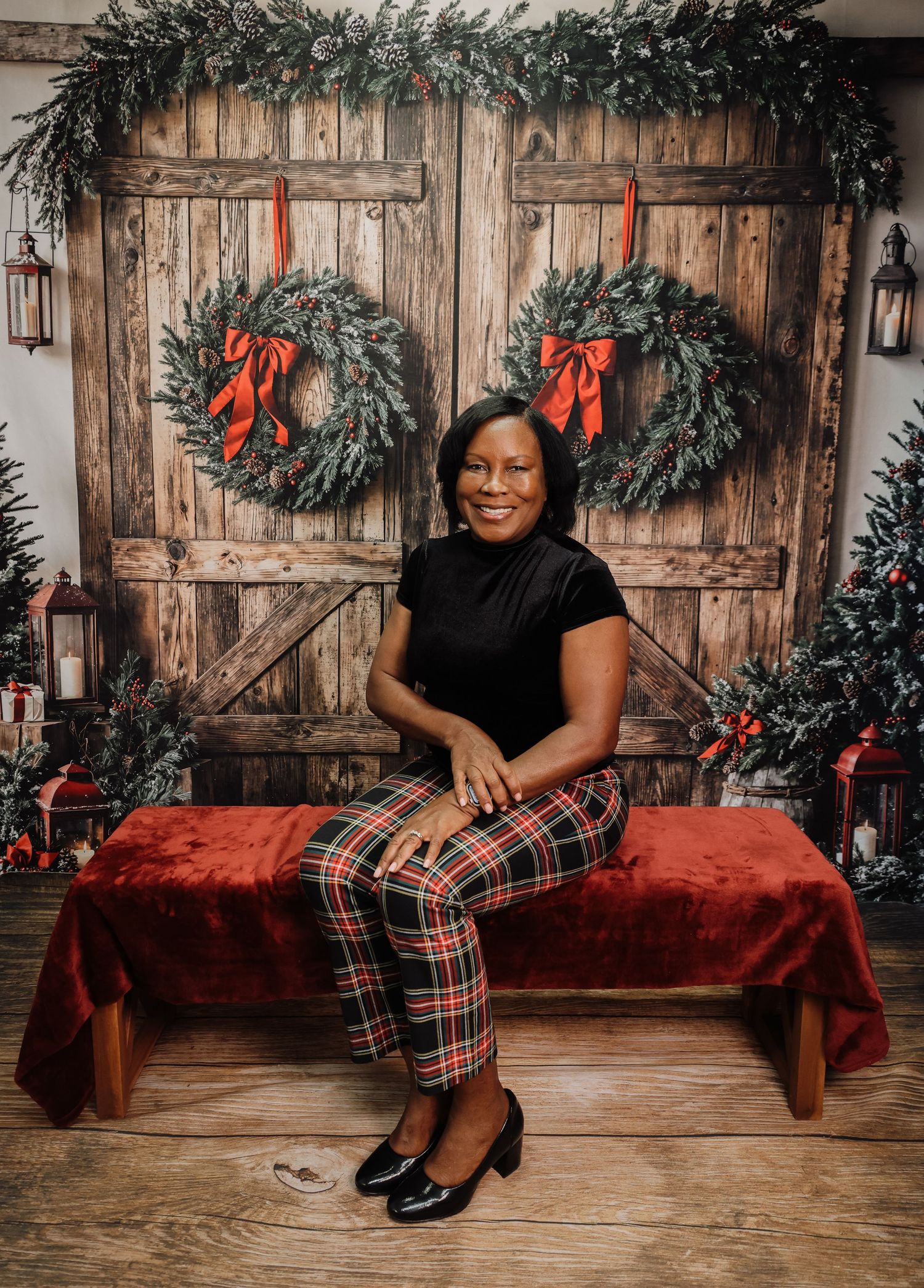 Woman in plaid pants sits on red bench before rustic barn doors adorned with Christmas wreaths and holiday decor.