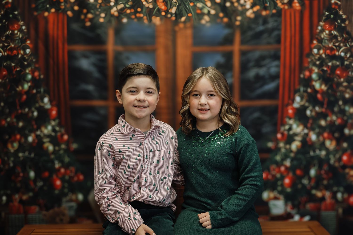 Two children pose together in front of decorated Christmas trees in a festive holiday portrait photo.