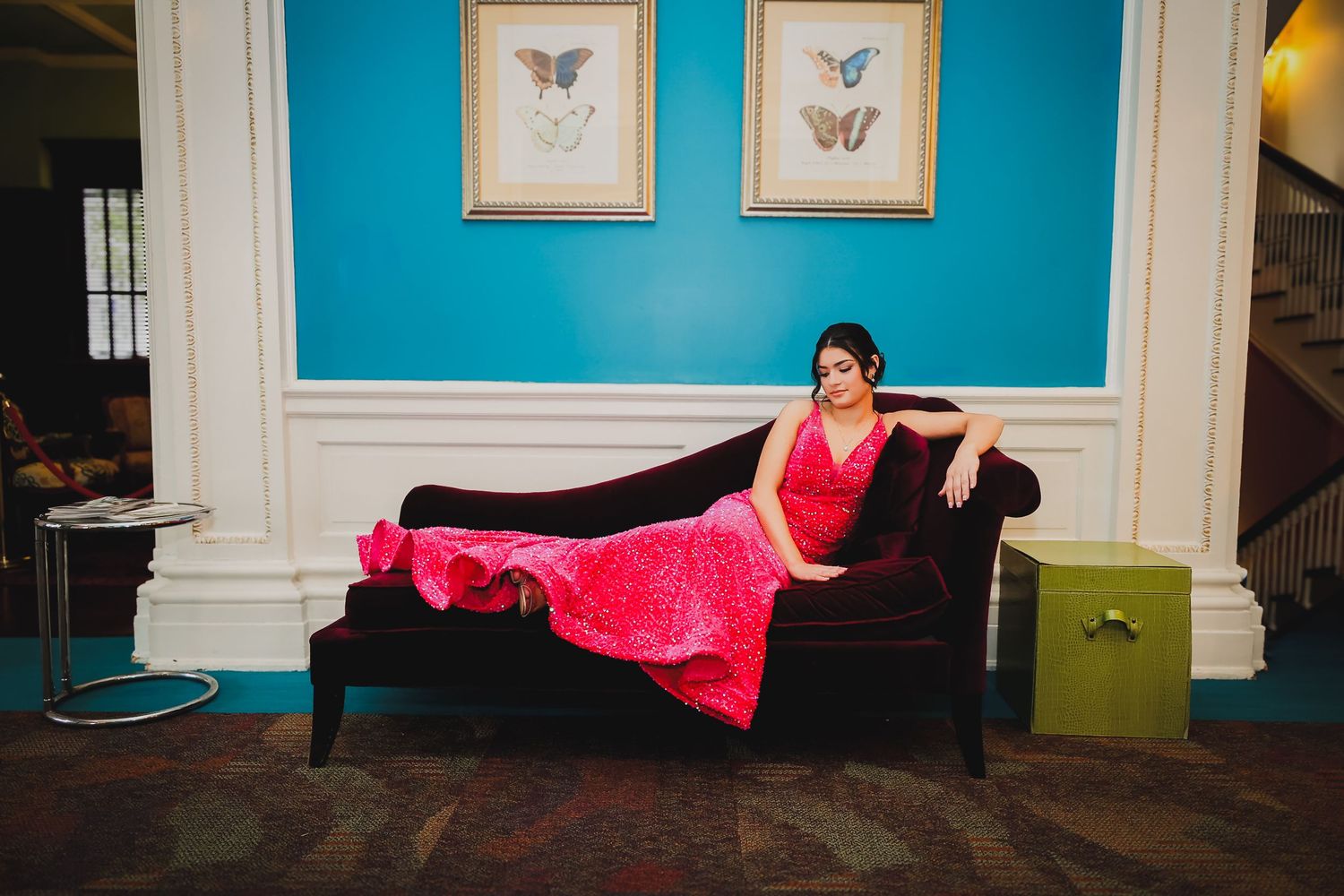 Woman in red gown reclining on dark velvet chaise lounge in elegant blue room with butterfly artwork.