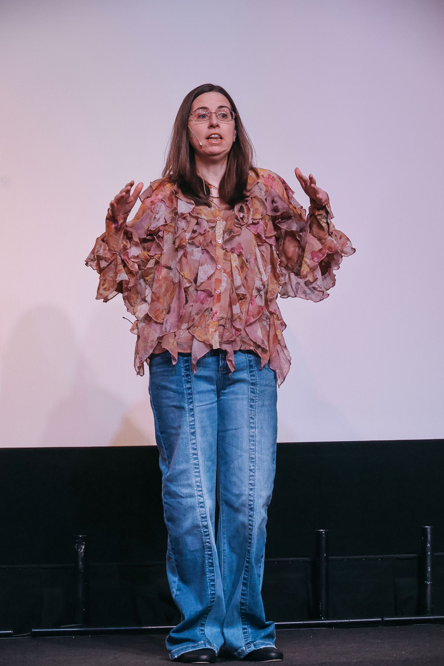 Woman in floral ruffled blouse and wide-leg jeans gesturing while speaking on stage.