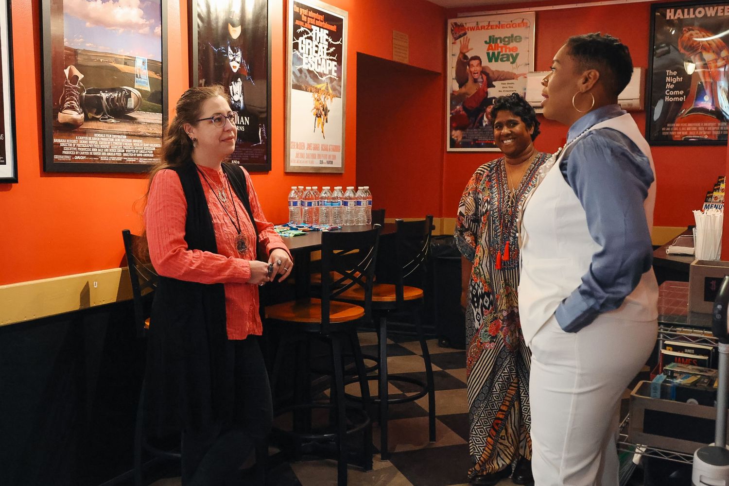 Three people conversing in a restaurant with orange walls and vintage movie posters, featuring casual and formal attire.