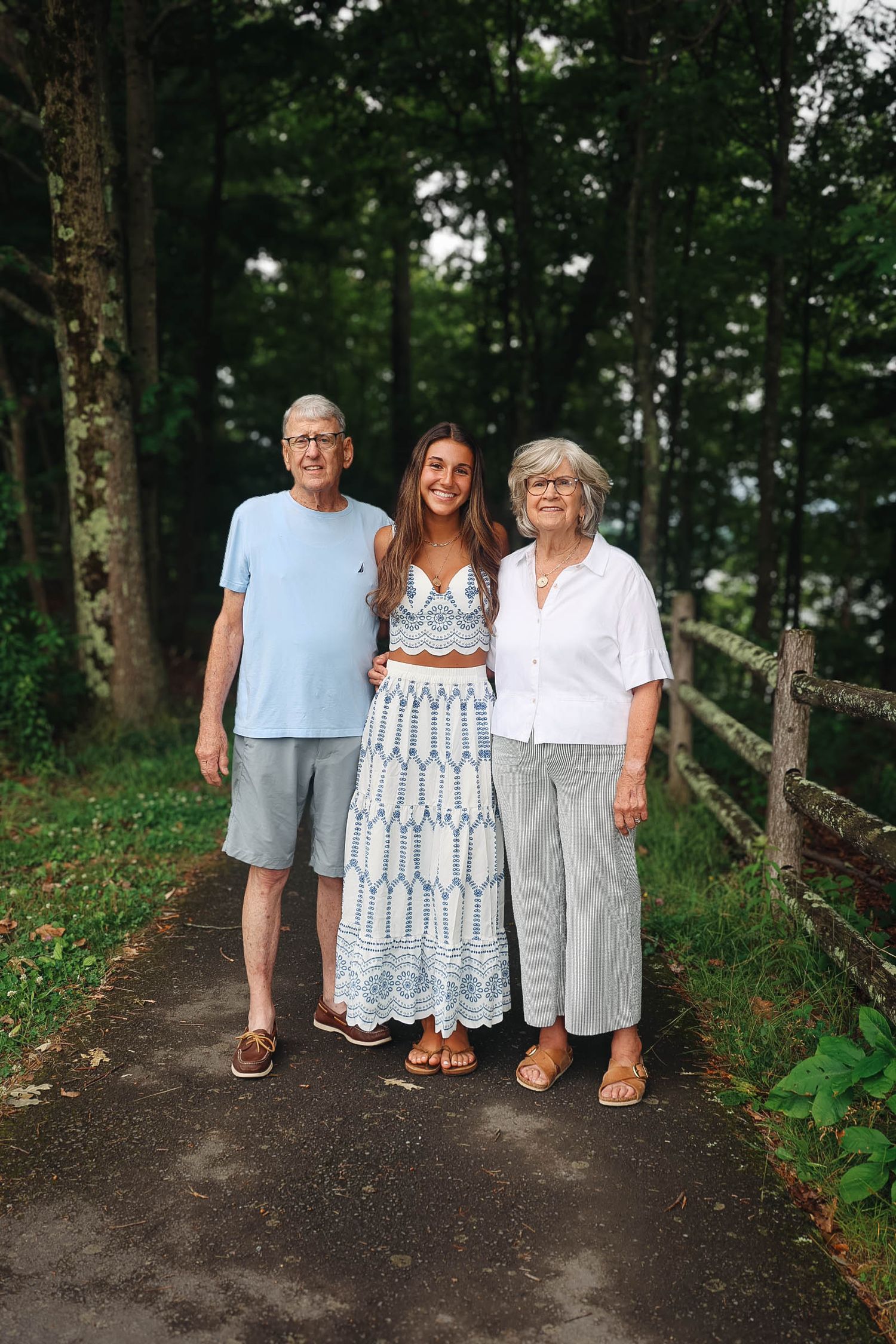 Young woman poses with elderly couple on a tree-lined path near a wooden fence on a summer day.