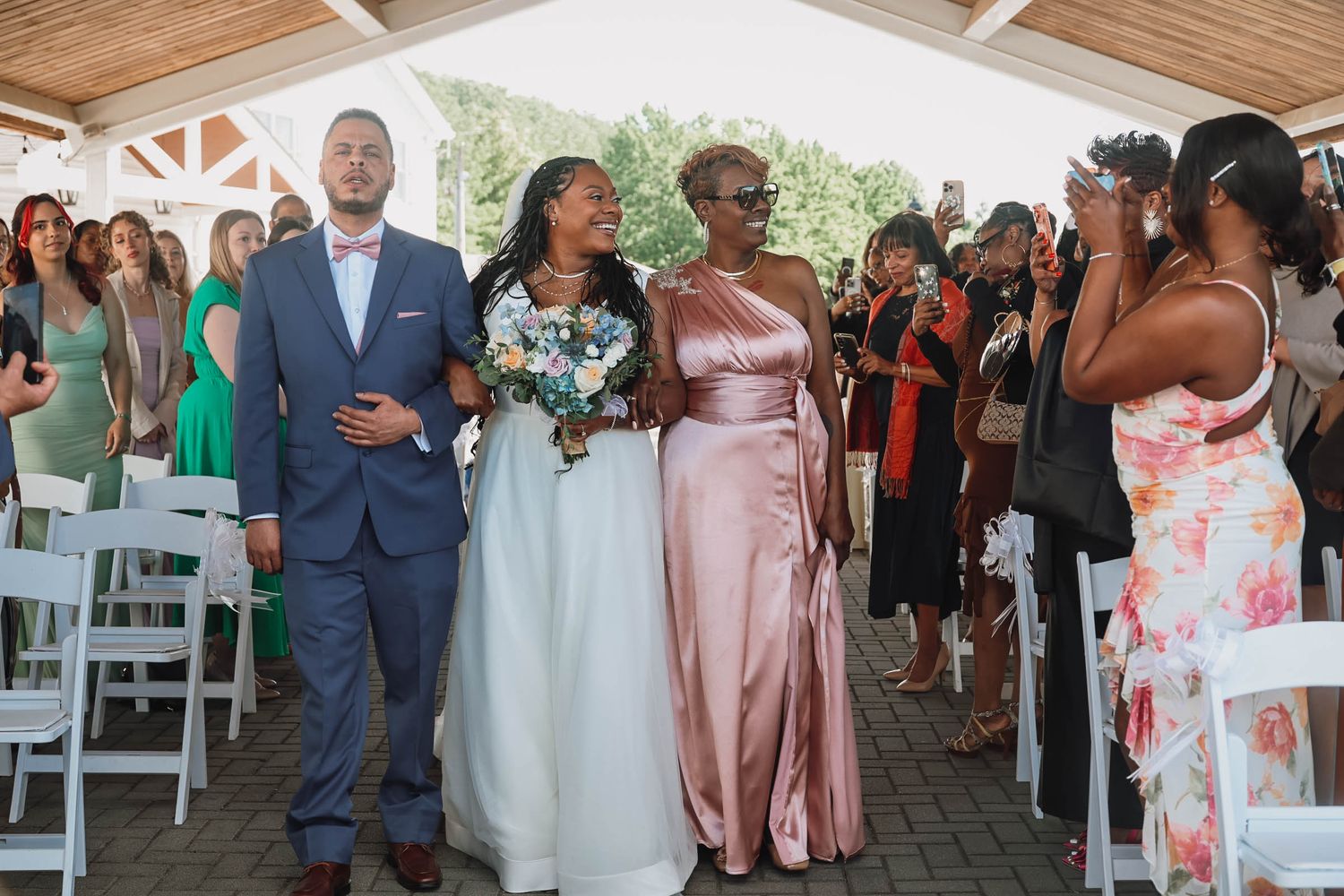 Bride walks down the aisle escorted by two loved ones at an outdoor wedding ceremony with seated guests.
