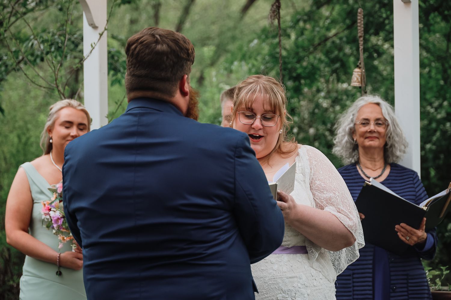 Bride reads wedding vows to groom during outdoor ceremony with officiant and bridesmaid in background.