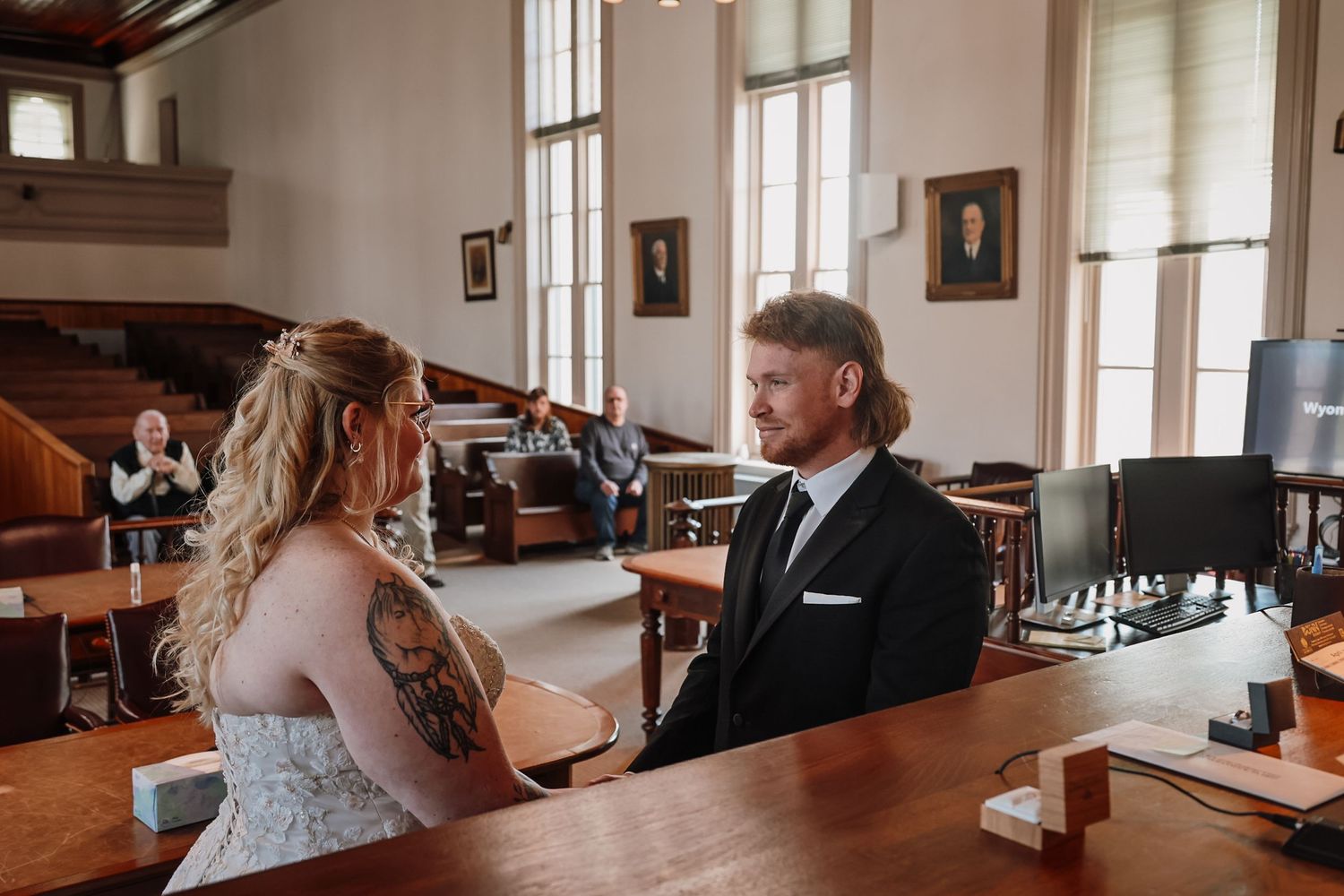 Couple exchanging vows at a courthouse wedding ceremony, bride with tattoos in white gown, groom in black suit.