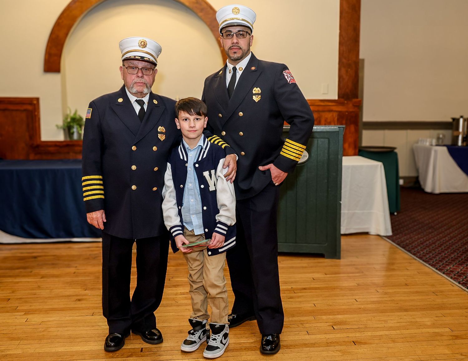 Two firefighters in dress uniforms pose with a young boy at a formal ceremony event.