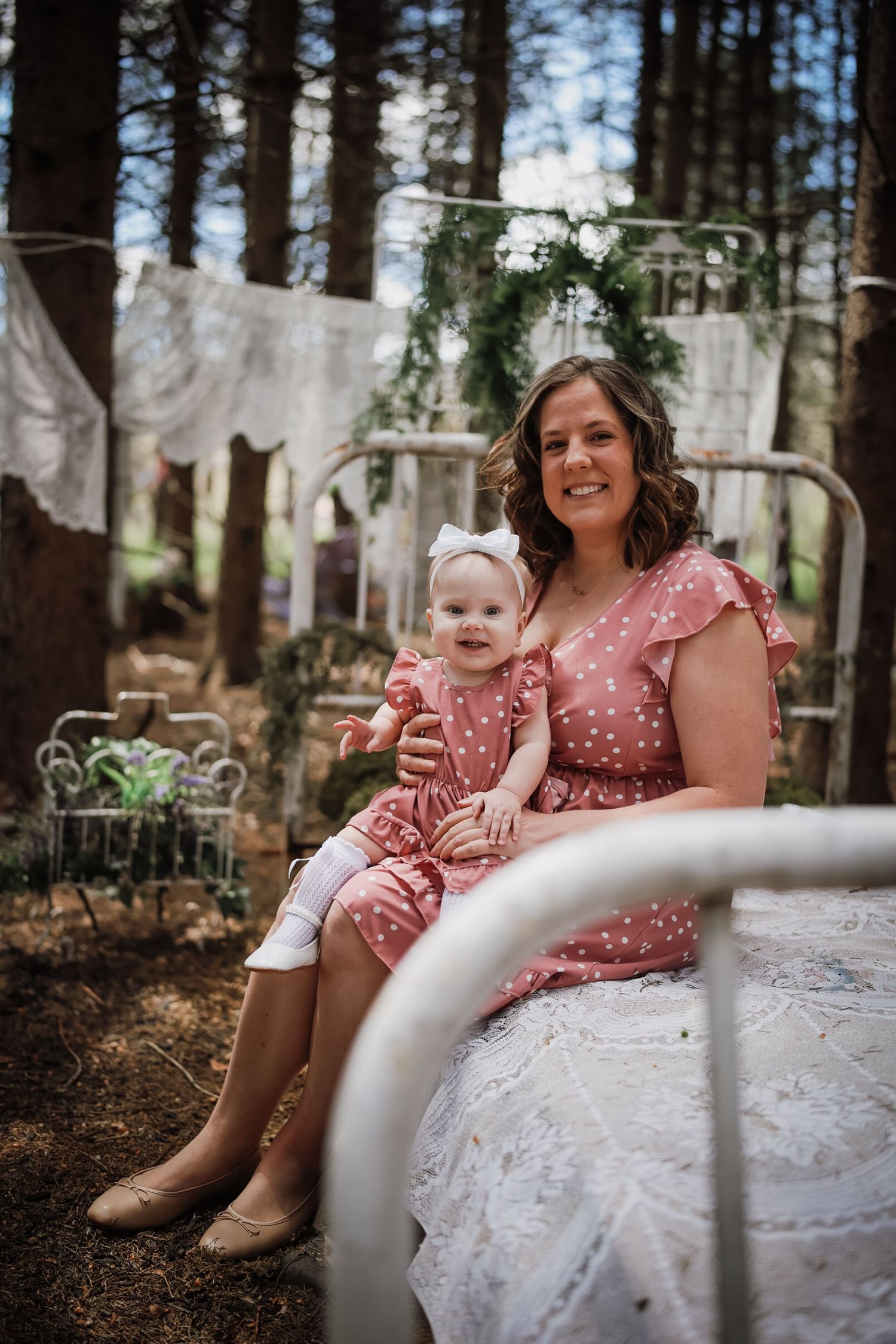 Mother and baby girl in matching pink polka dot dresses sitting on white iron bed in enchanted forest setting.