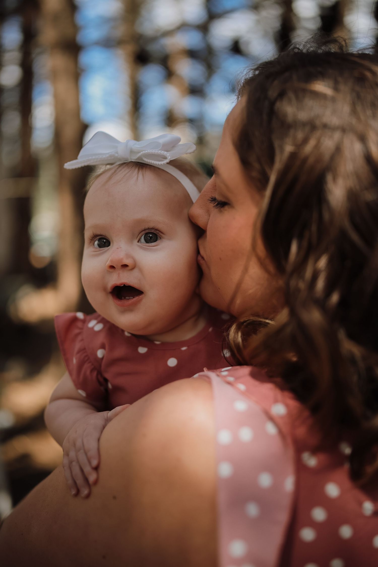 A mother kissing her happy baby girl wearing a white bow and pink polka dot dress outdoors in a forest.