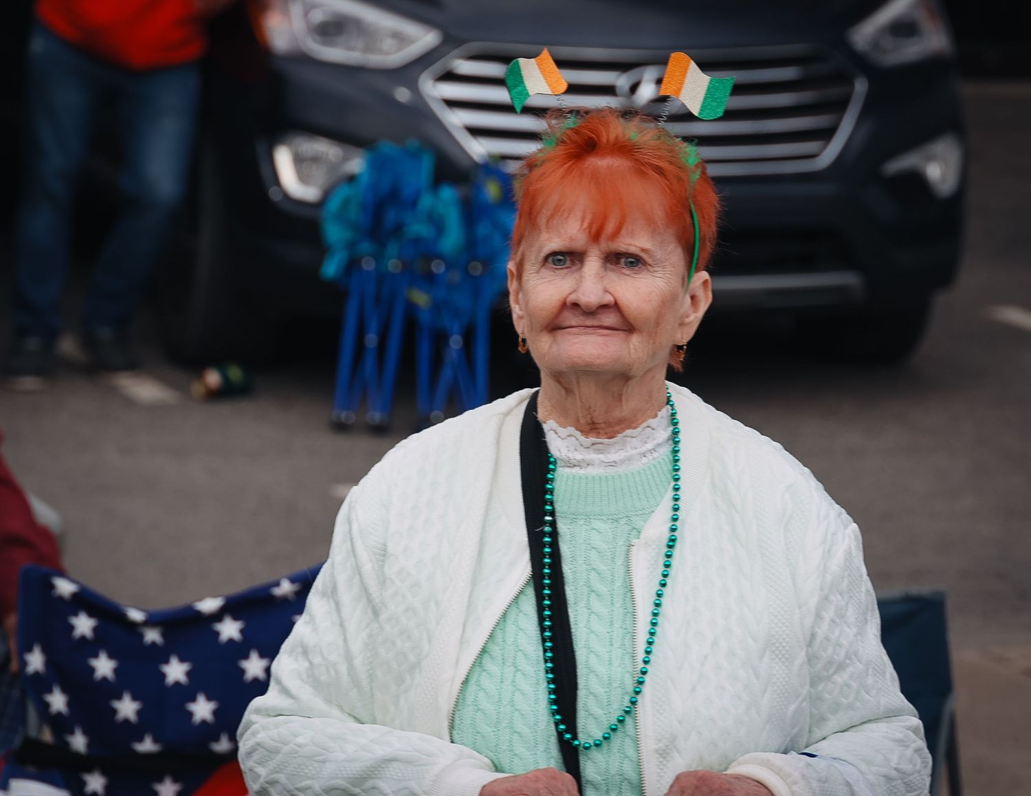 Woman with orange hair wearing green beads and Irish flag headband at St. Patrick's Day parade.