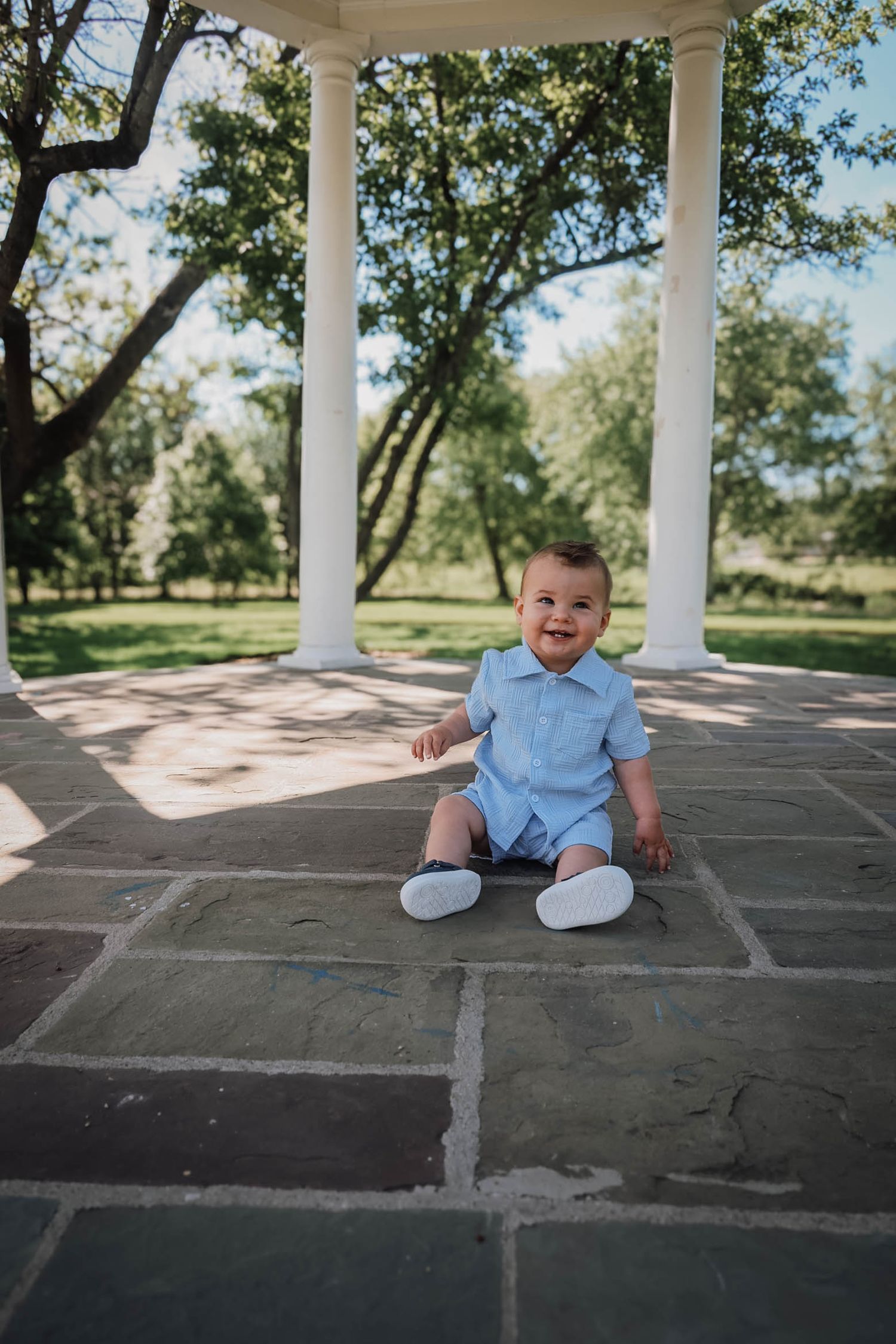 A smiling baby boy in a blue outfit sits on stone pavers under a white columned gazebo surrounded by green trees.