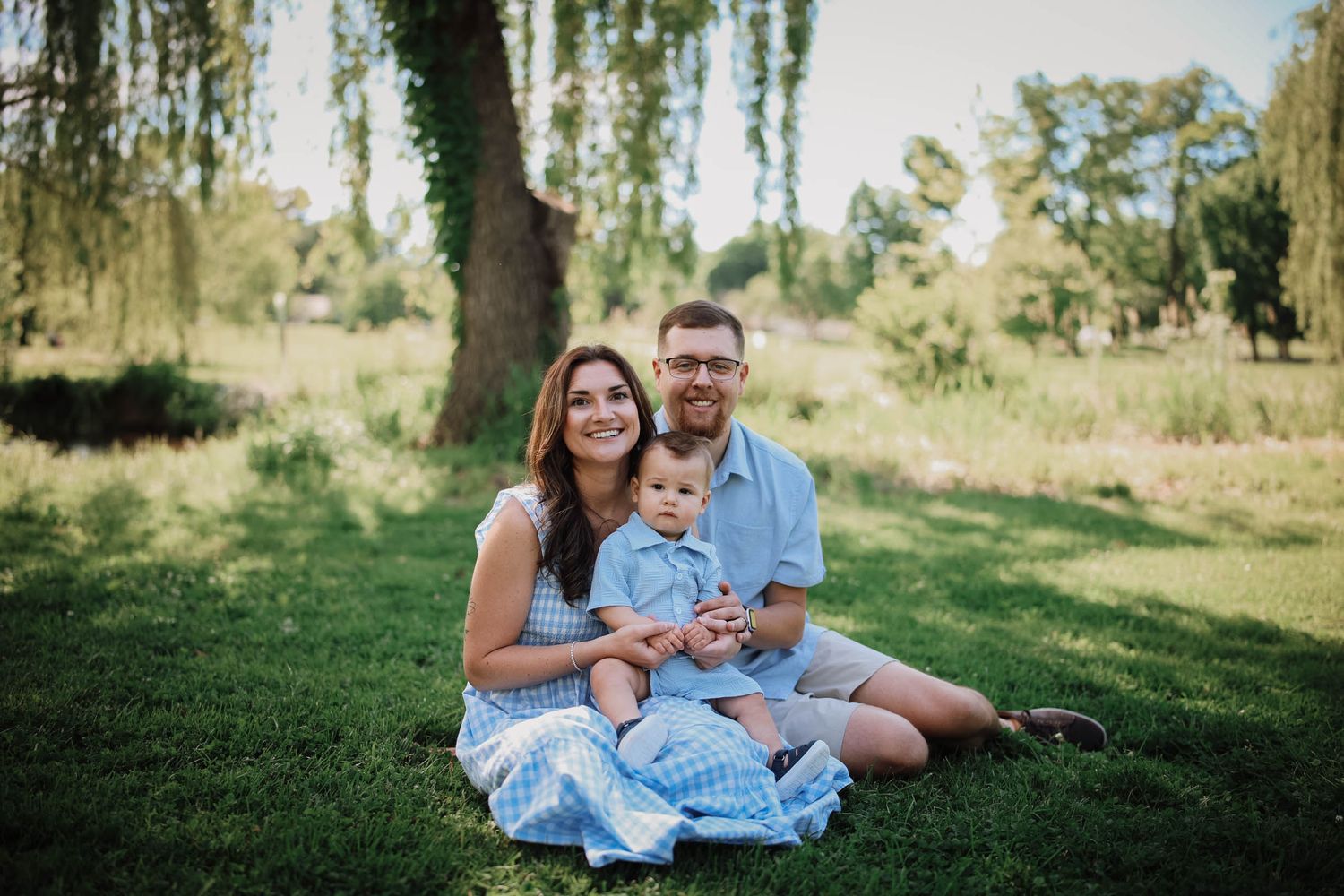 A happy family of three sits on green grass under a willow tree, parents smiling with their baby boy outdoors.