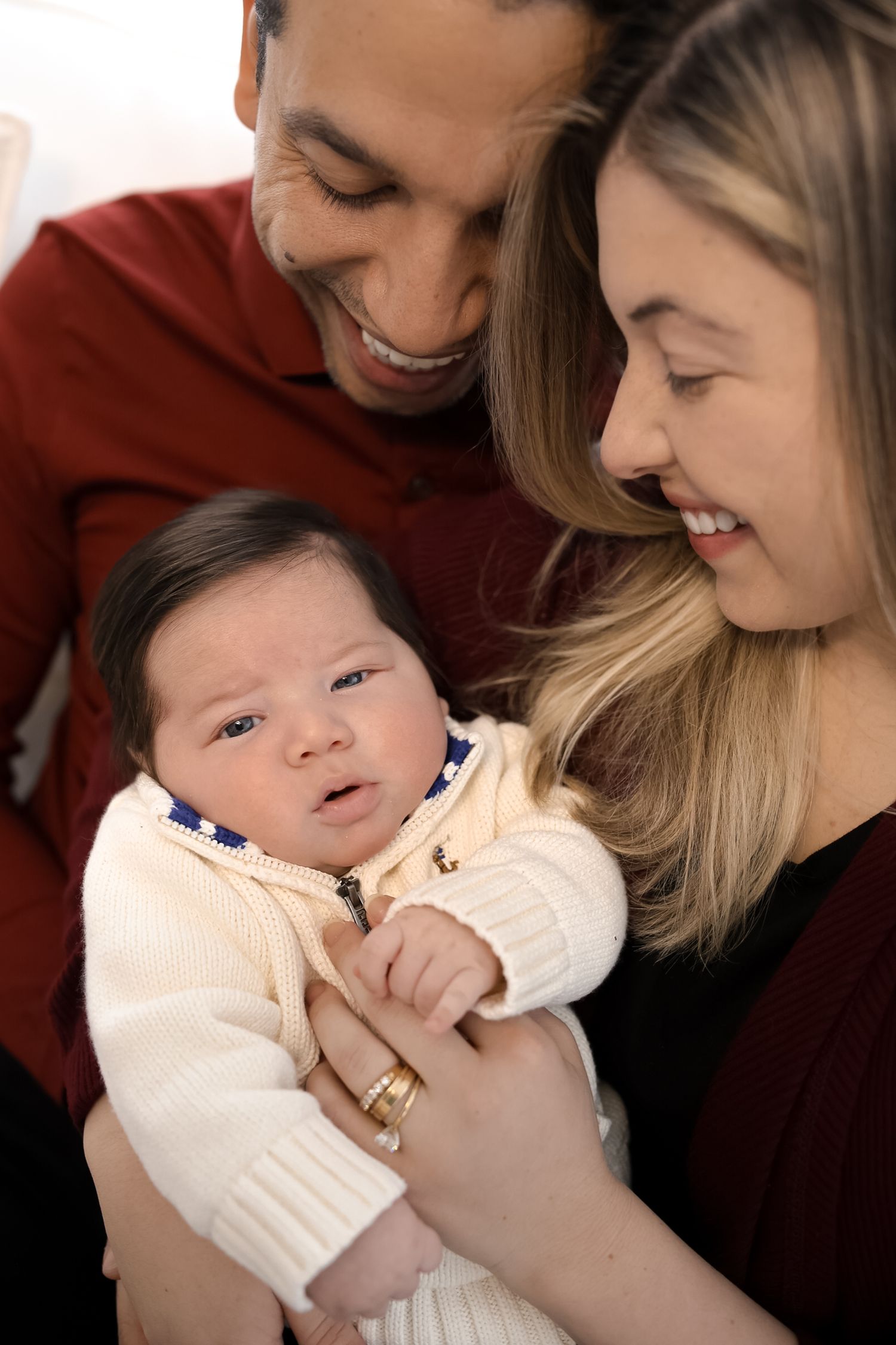 A tender moment captured between parent and baby in white clothing against a warm burgundy background.