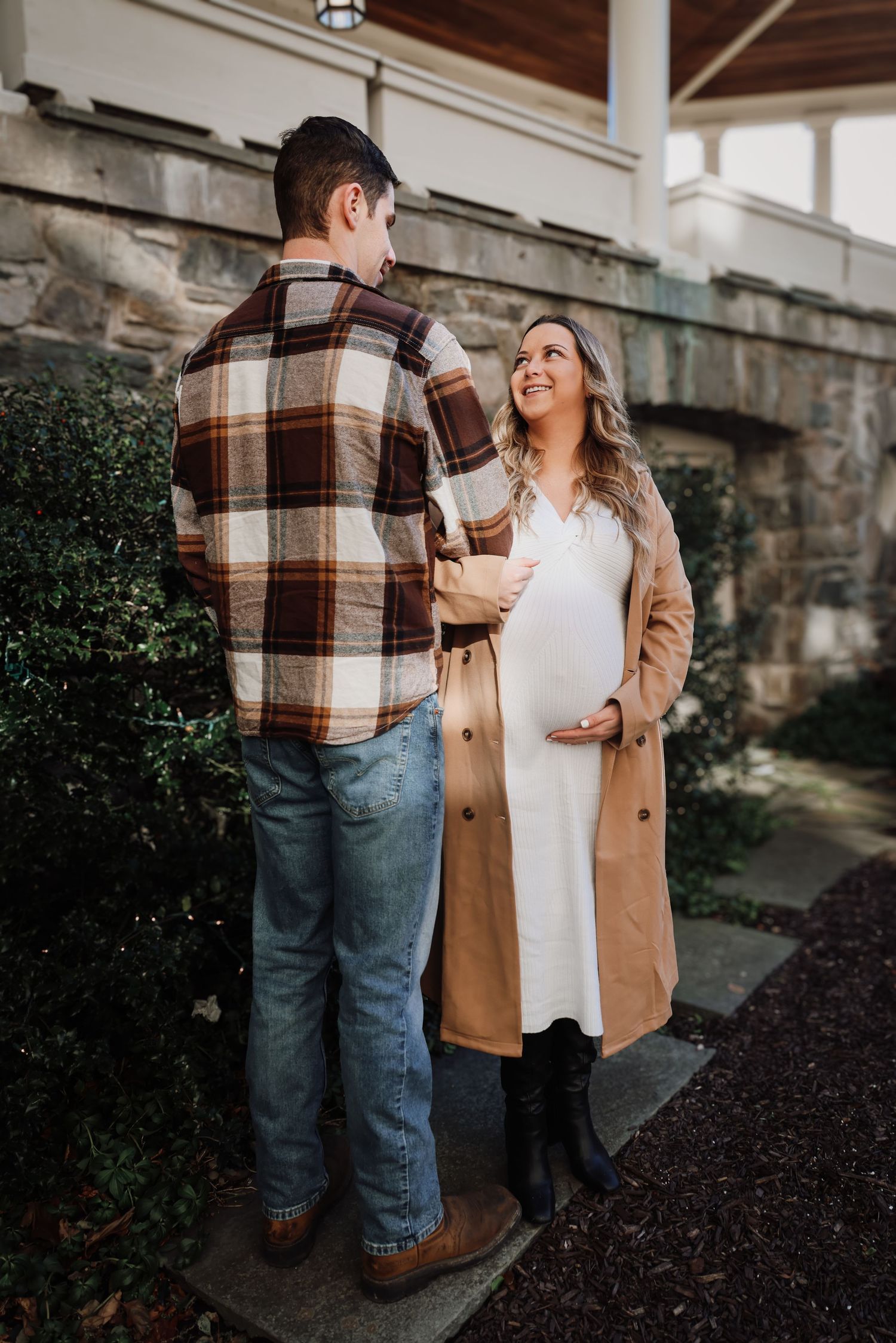 A couple in casual fall attire shares a tender moment outdoors near a stone wall and garden.