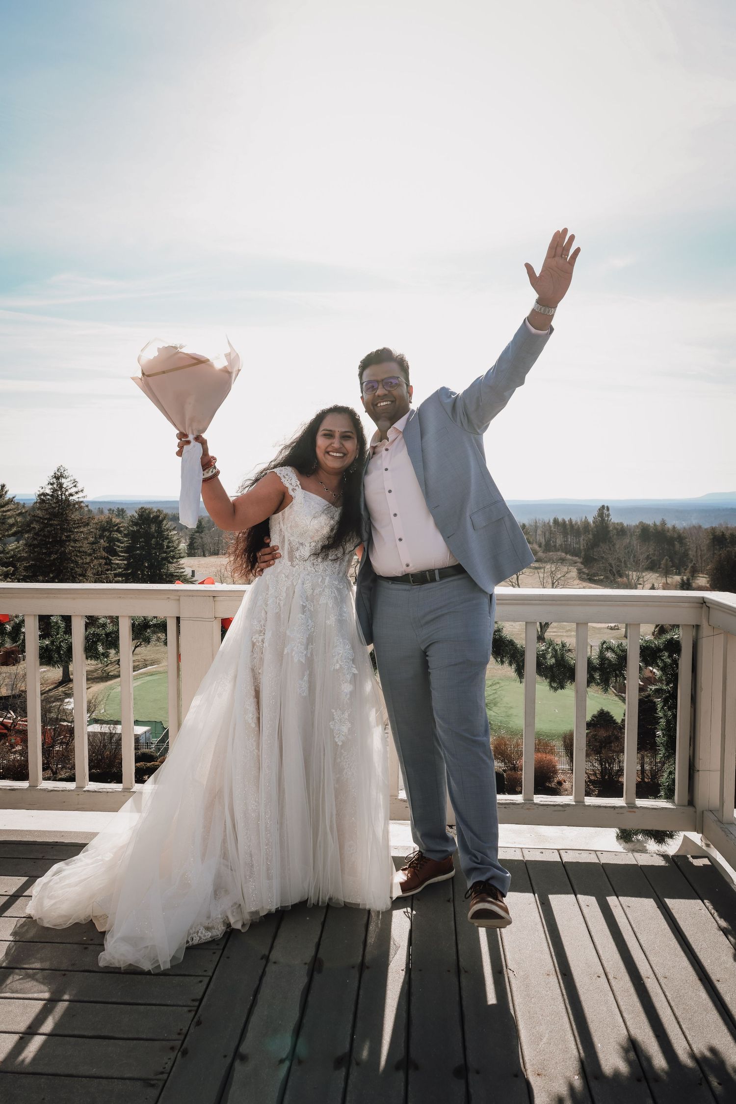 A couple in wedding attire celebrates joyfully on a wooden deck overlooking a scenic mountain landscape.