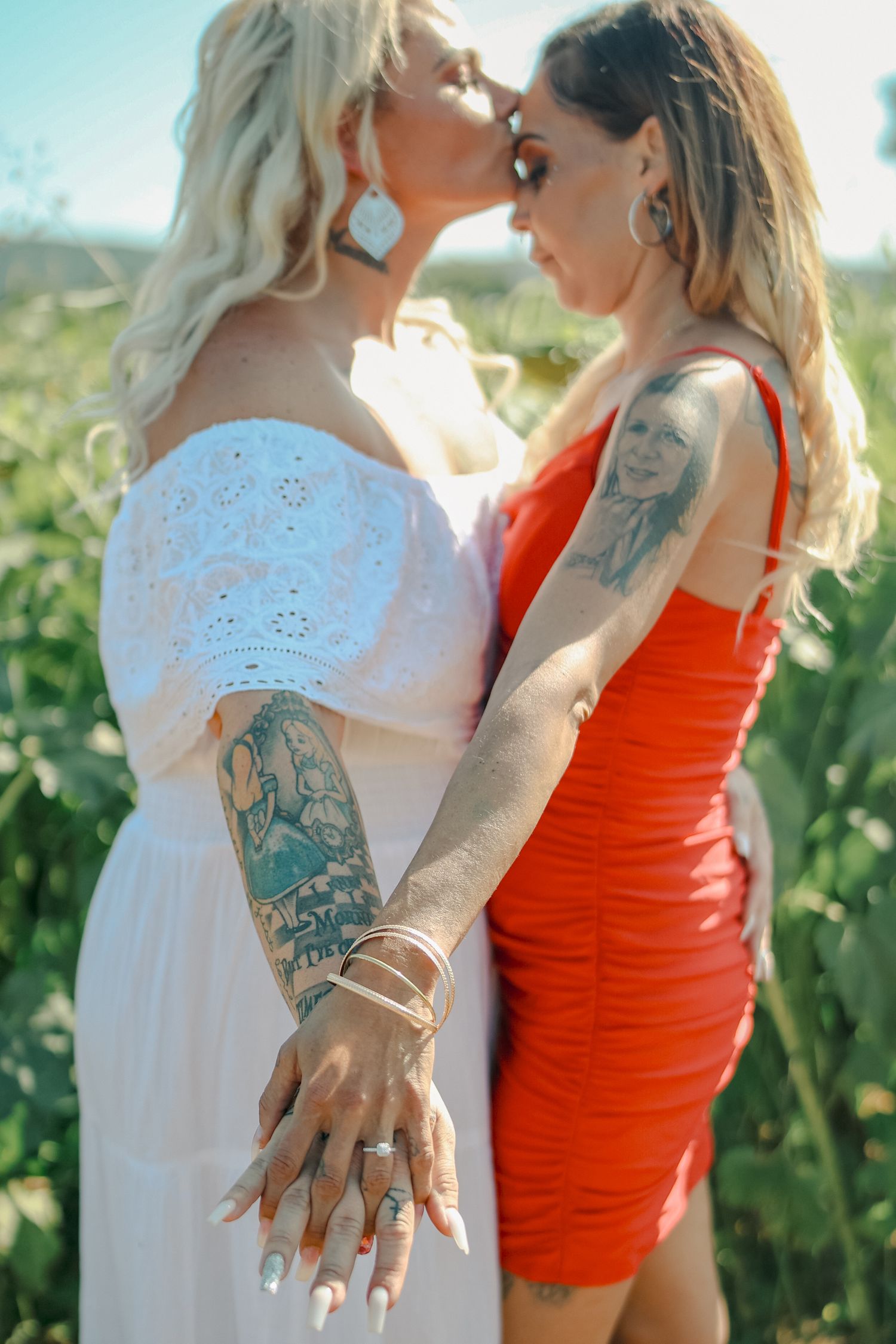 Two people in white and orange outfits stand close together in a vineyard setting showing rings and tattoos.