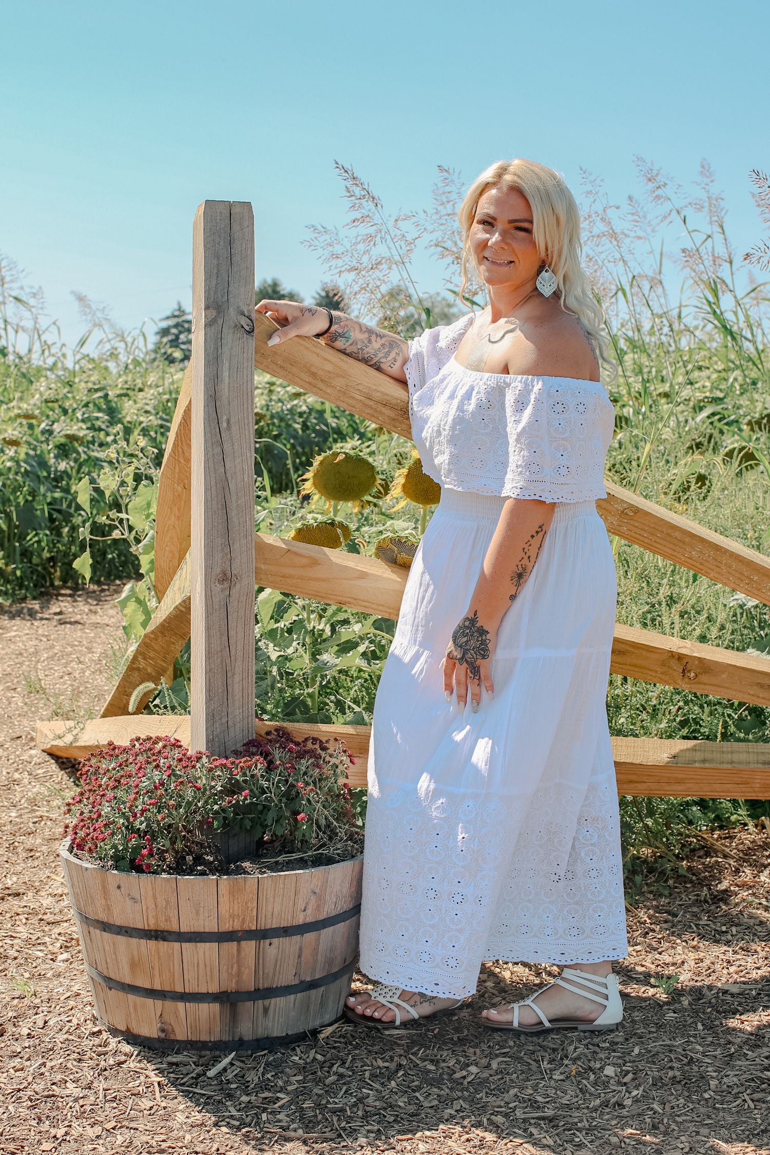 Woman in white eyelet dress poses near rustic wooden fence and barrel planters in summer garden setting.