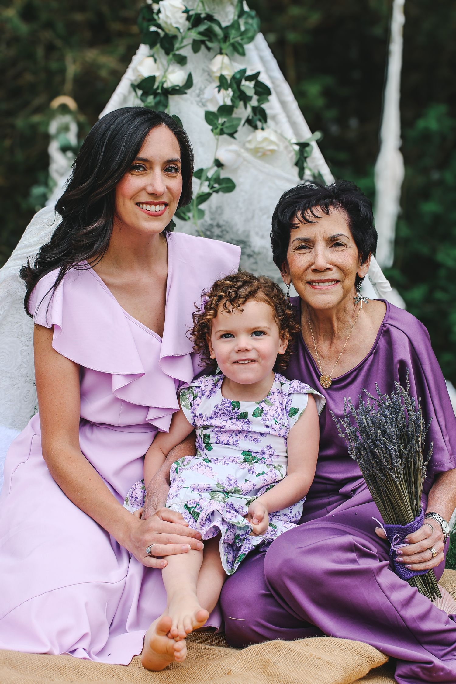 Three generations of family members wearing pink and purple dresses pose together in a garden setting.