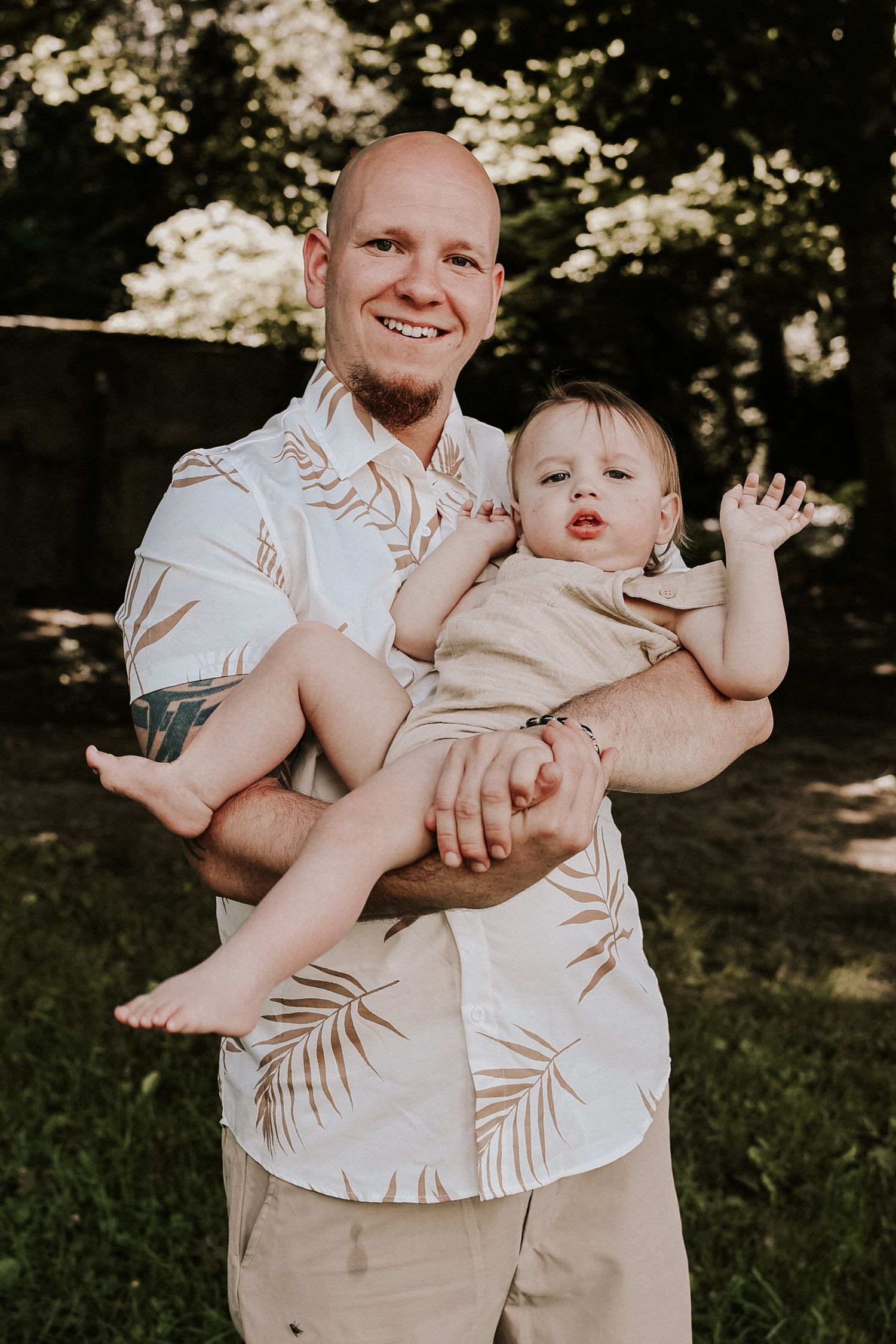 A parent and child wearing matching palm print shirts pose together outdoors in dappled sunlight.