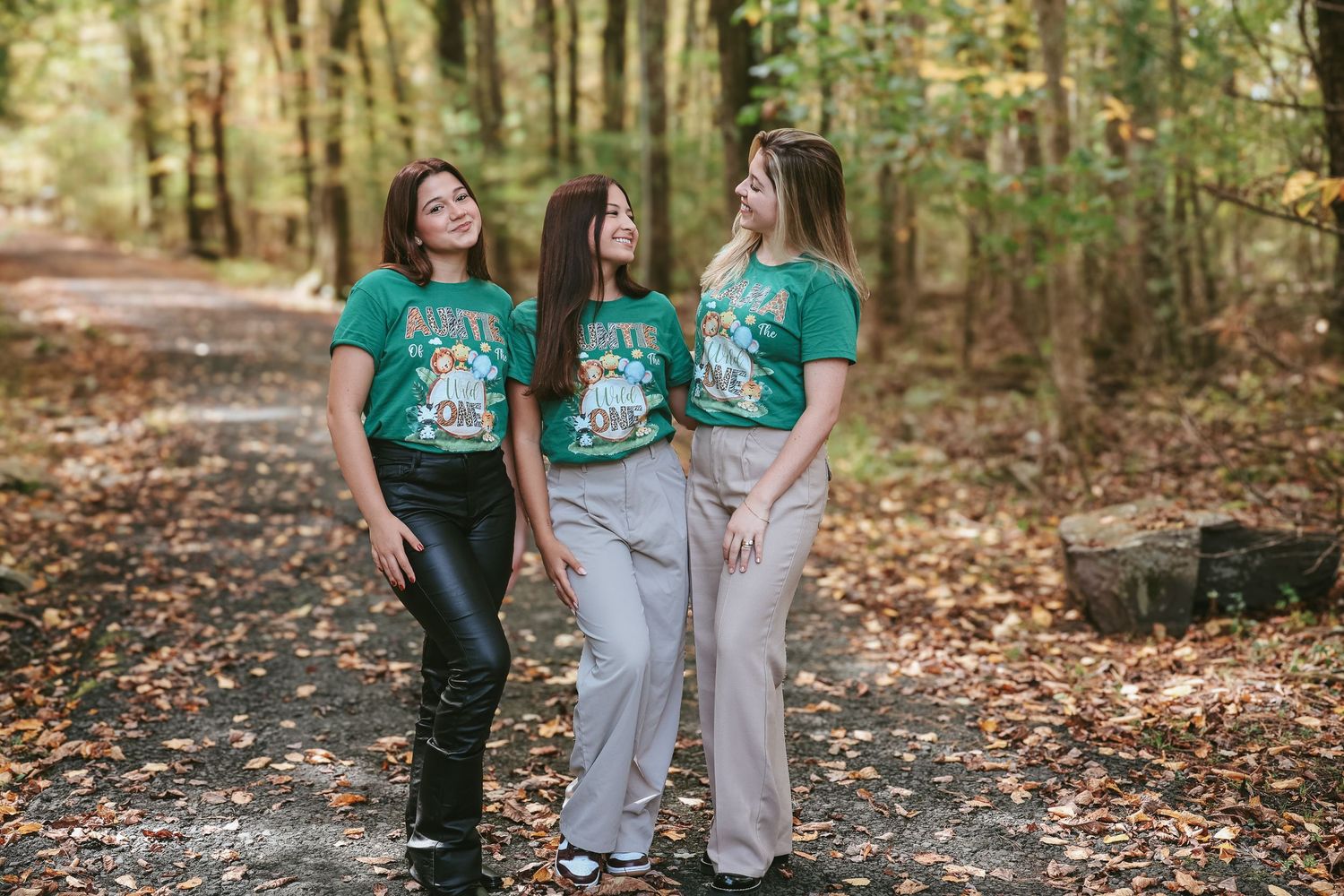 Three people wearing matching green and gray outfits stand together in a wooded area during autumn.