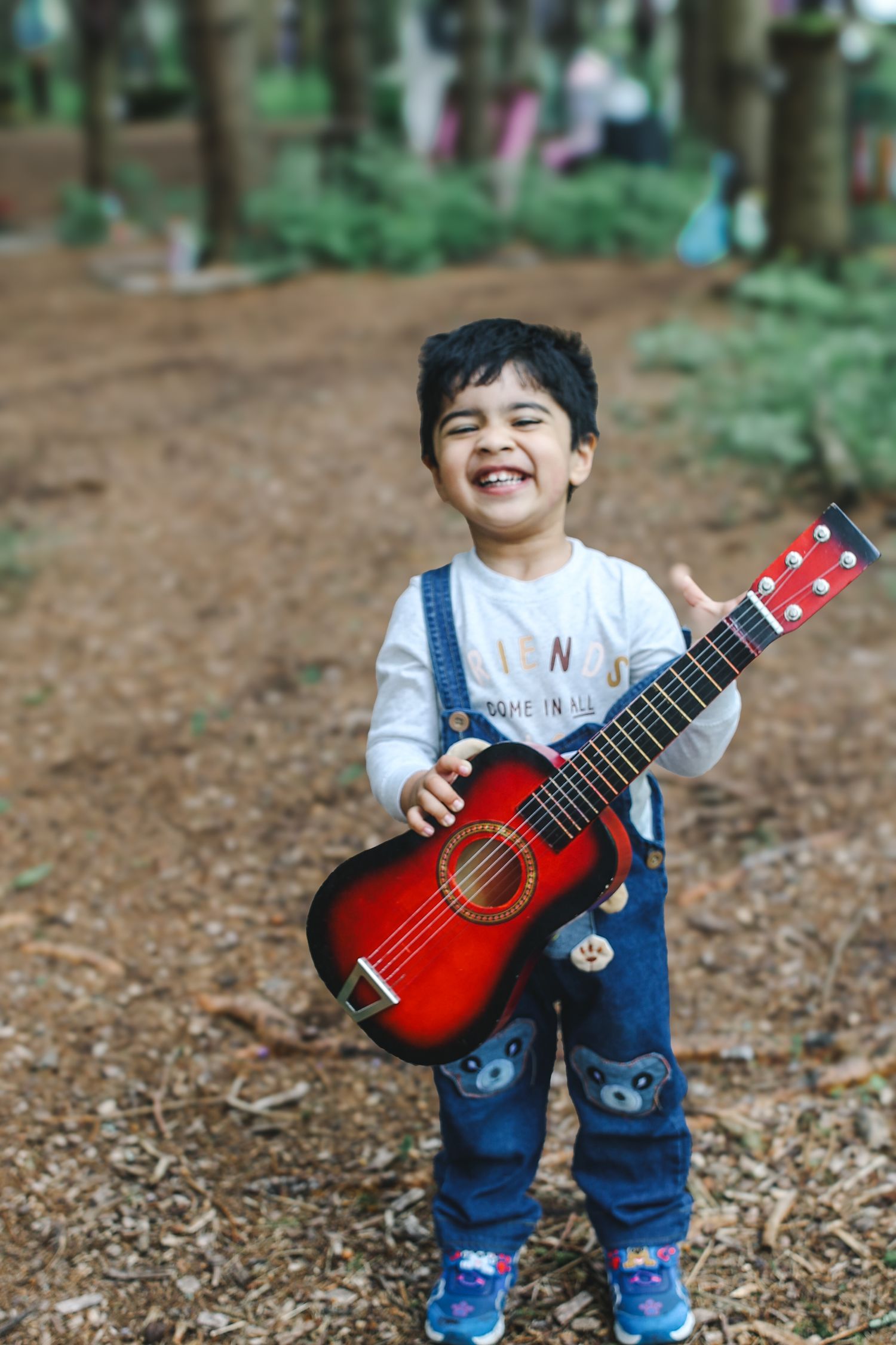 Young child in denim overalls smiles while holding a red acoustic guitar in a wooded outdoor setting.