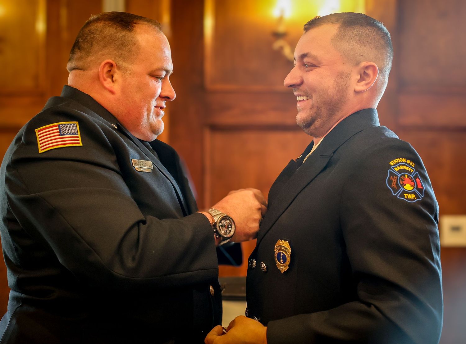Two people in suits share a warm embrace and laughter in a warmly lit indoor setting.