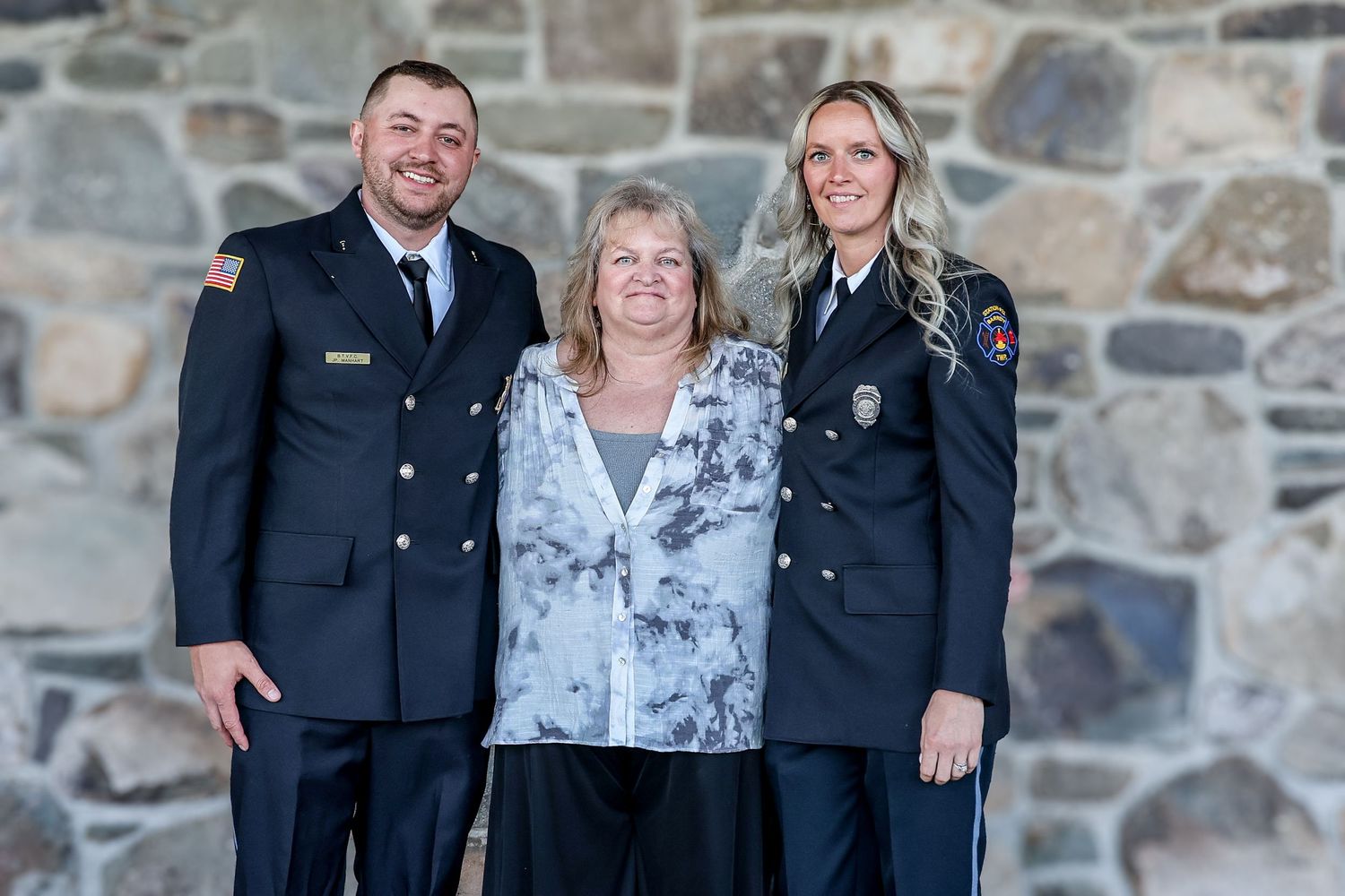 Three people pose together against a stone wall, with two wearing dark uniforms and one in a light patterned blouse.