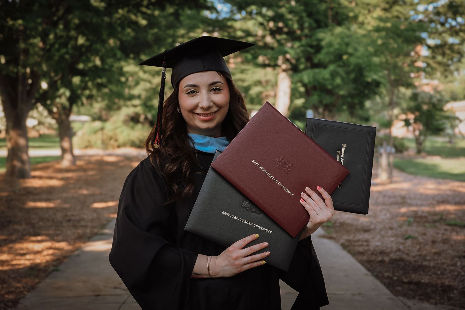 Graduate in cap and gown holds diploma while smiling outdoors on campus during autumn.