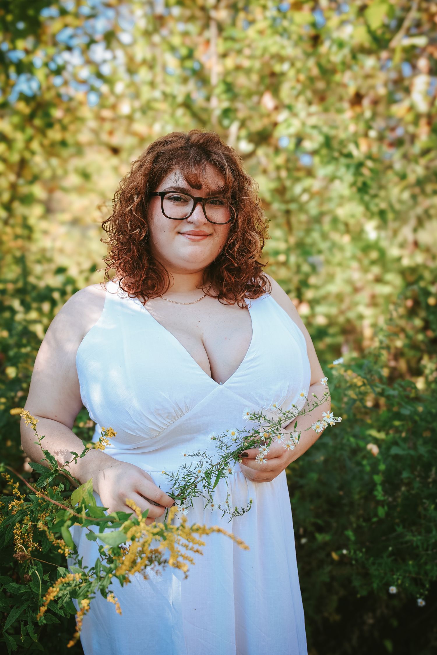 A person in a flowing white dress poses outdoors against lush green foliage on a sunny day.