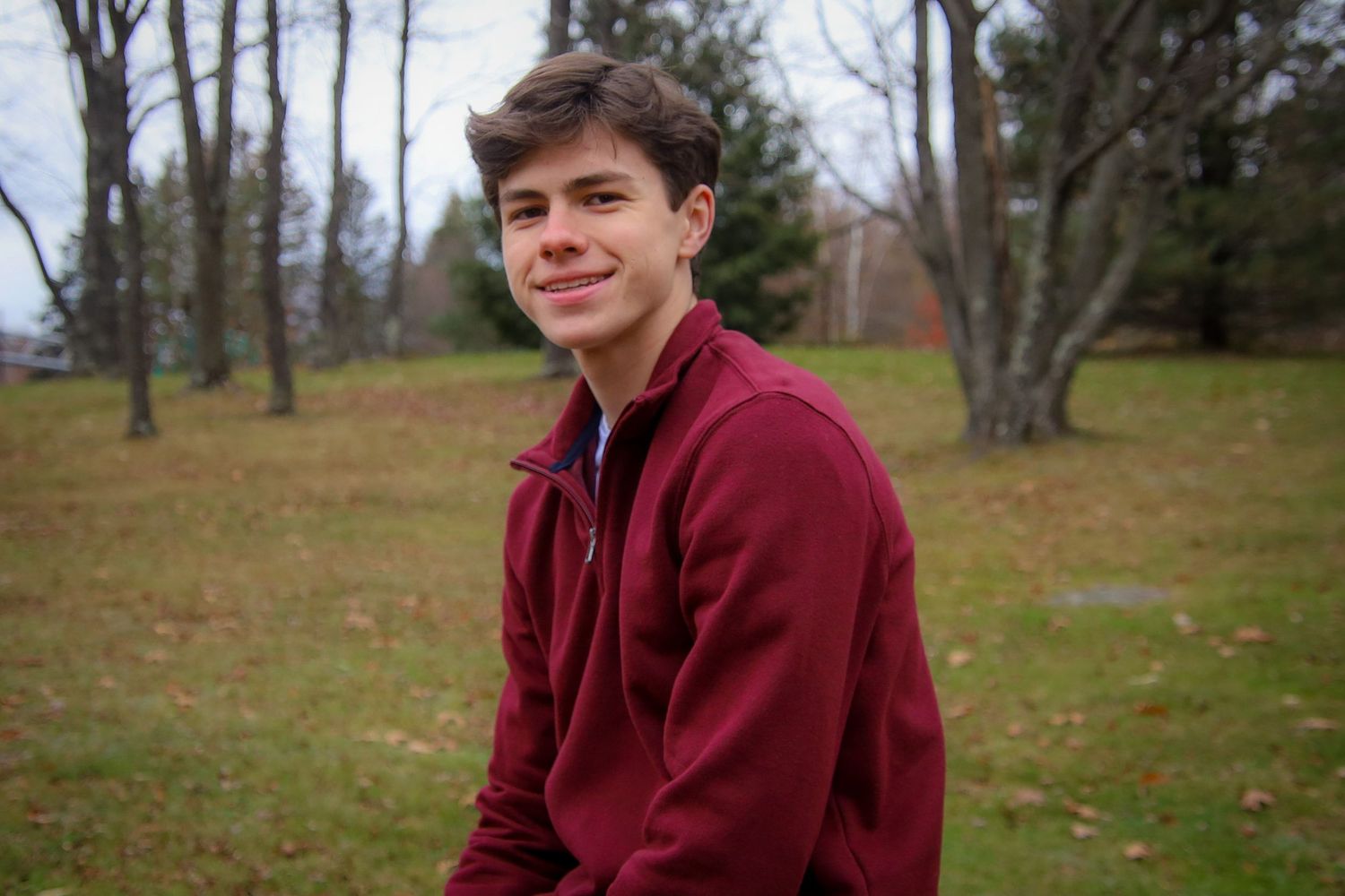 A person in a burgundy hoodie sits outdoors on grassy terrain surrounded by bare trees on an autumn day.