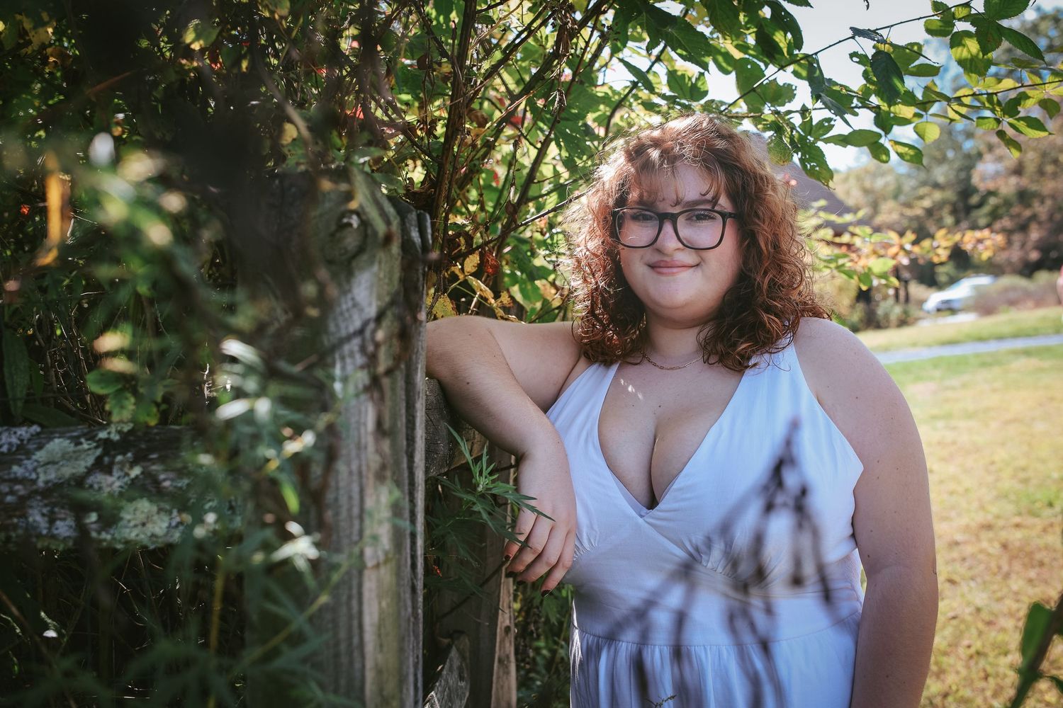 Woman in white dress poses in outdoor garden setting with natural greenery and foliage in background.
