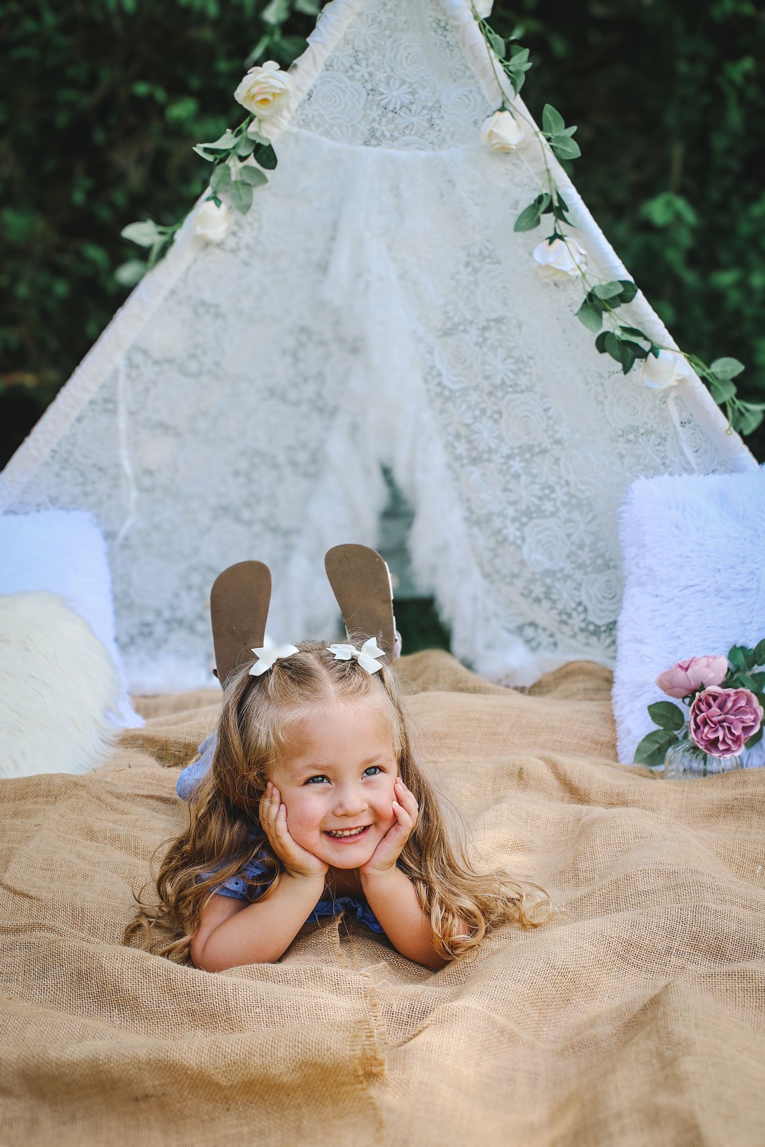 A child lies on a tan blanket in front of a white lace teepee decorated with roses and greenery.