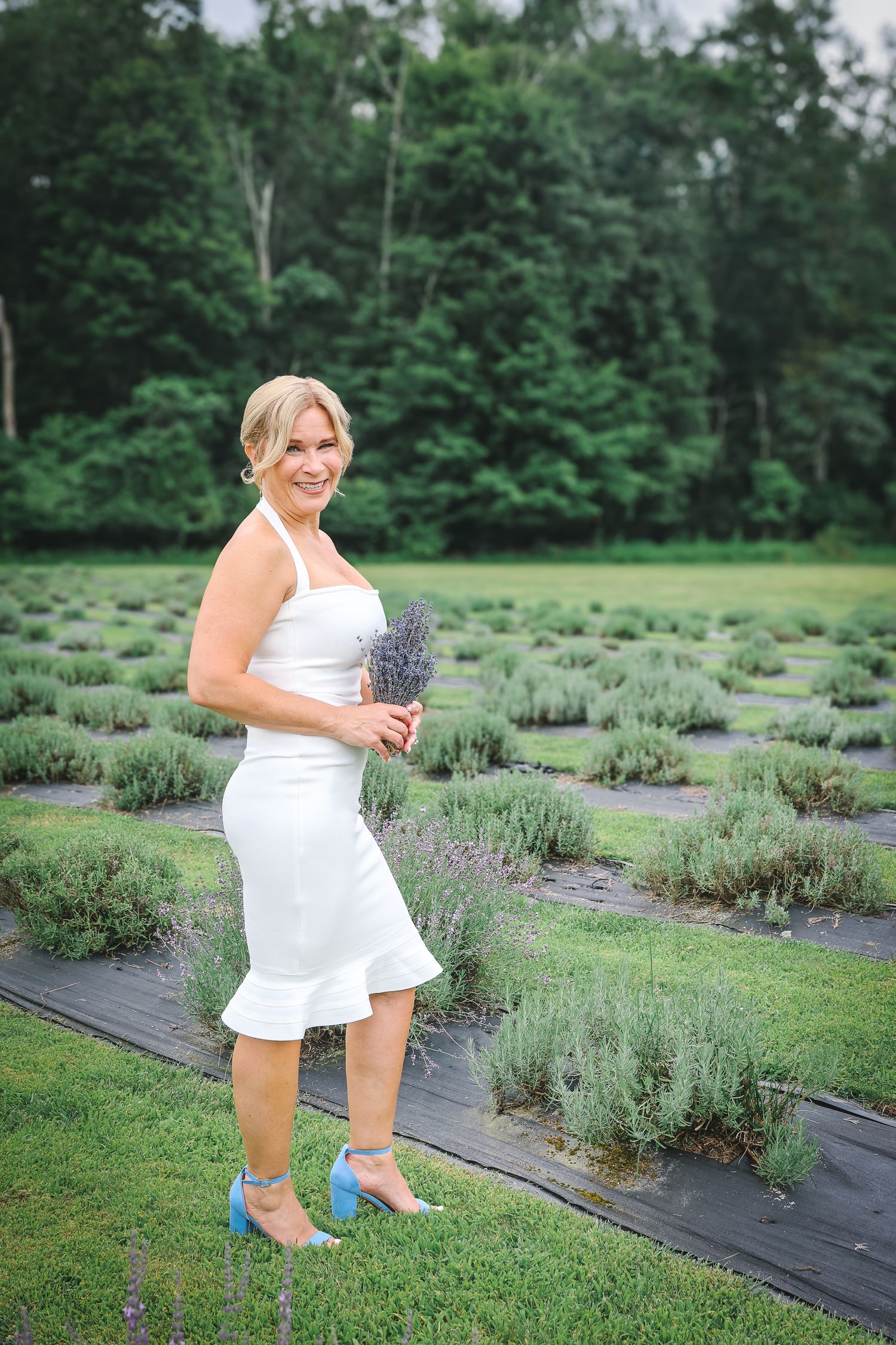 A person in a white dress poses in a lavender field with rows of purple flowers on a summer day.