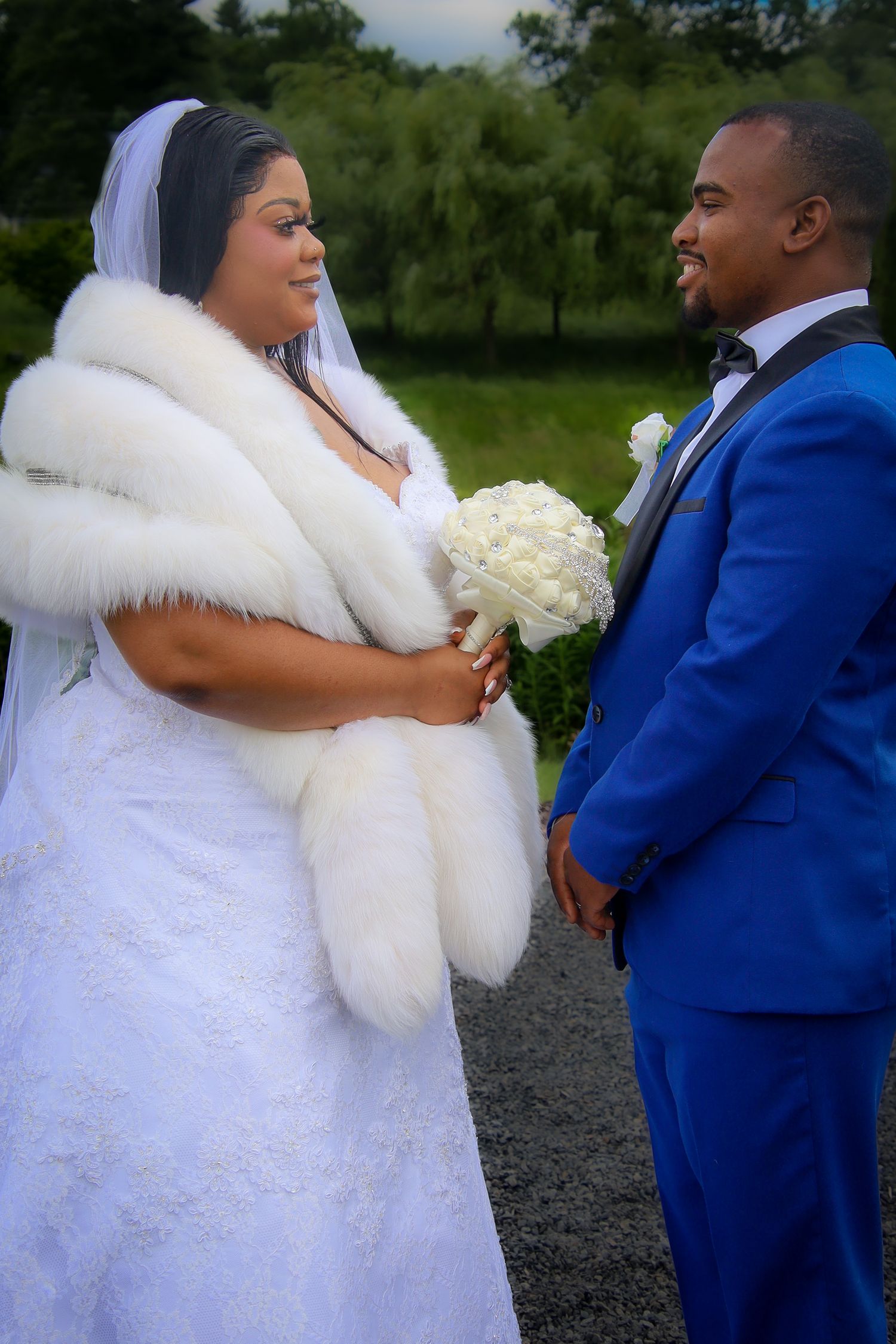 A couple in wedding attire, with the bride in white fur and lace gown and groom in royal blue suit, share intimate moments outdoors.