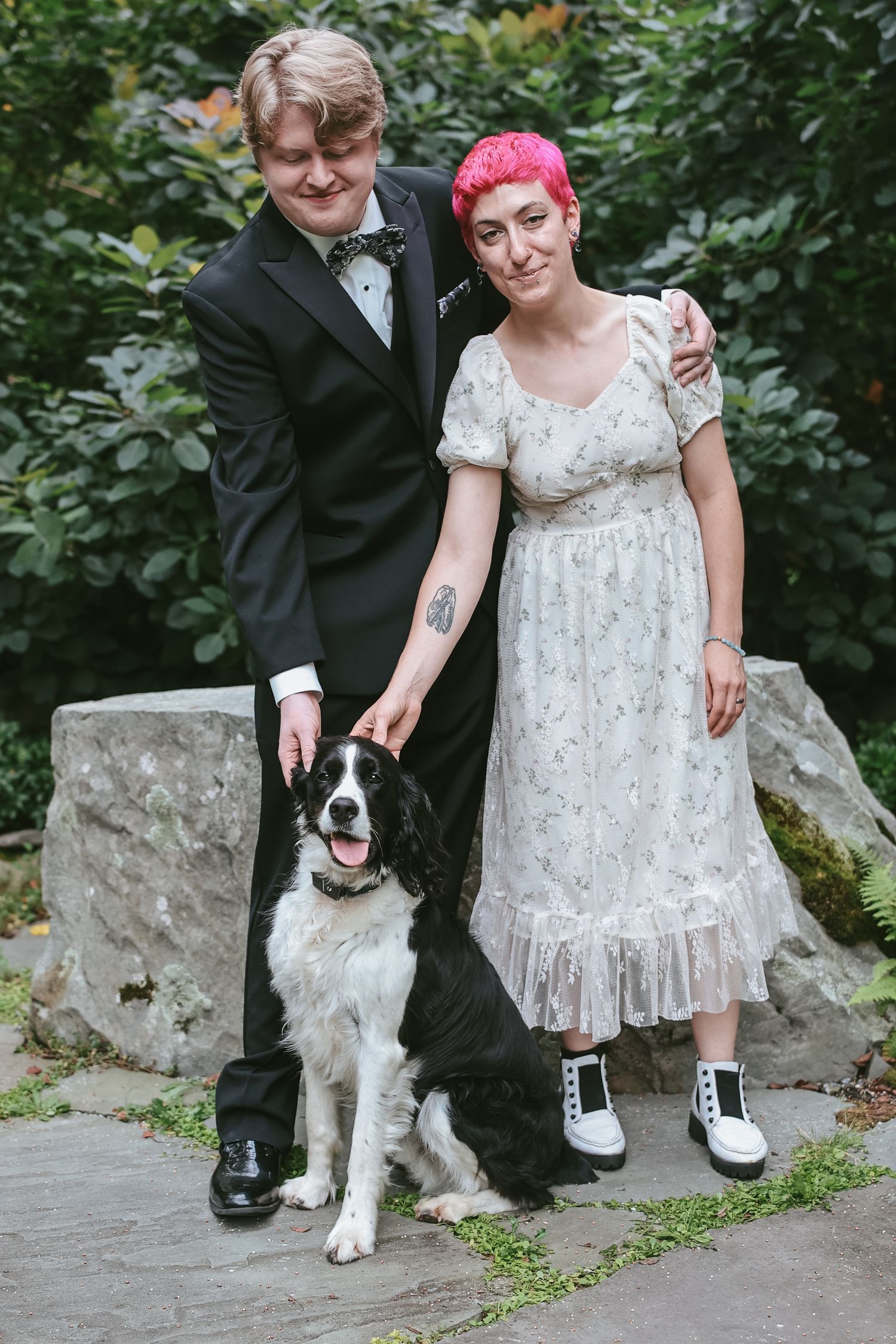 A couple in wedding attire poses with their black and white dog on stone steps surrounded by greenery.
