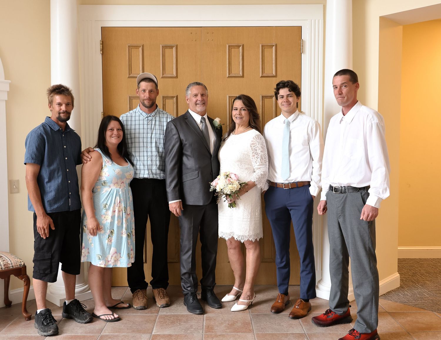 A wedding party stands in a row against a beige wall wearing formal attire and holding white bouquets.