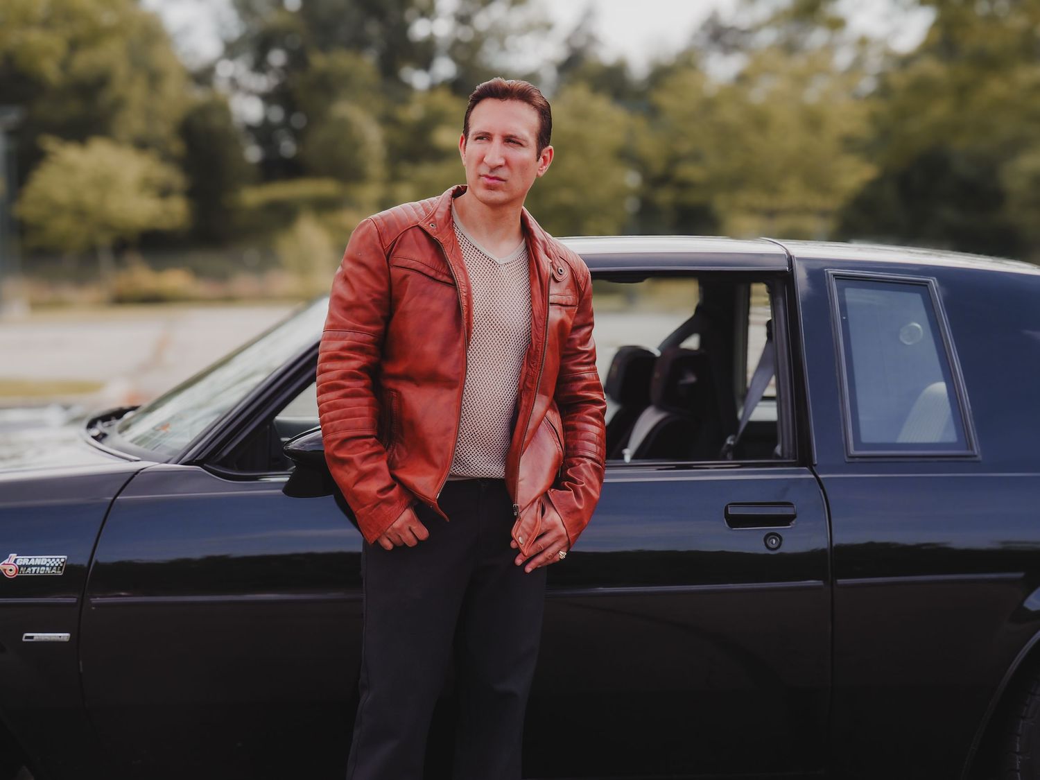 Someone in a red leather jacket leans against a black car in a wooded outdoor setting.
