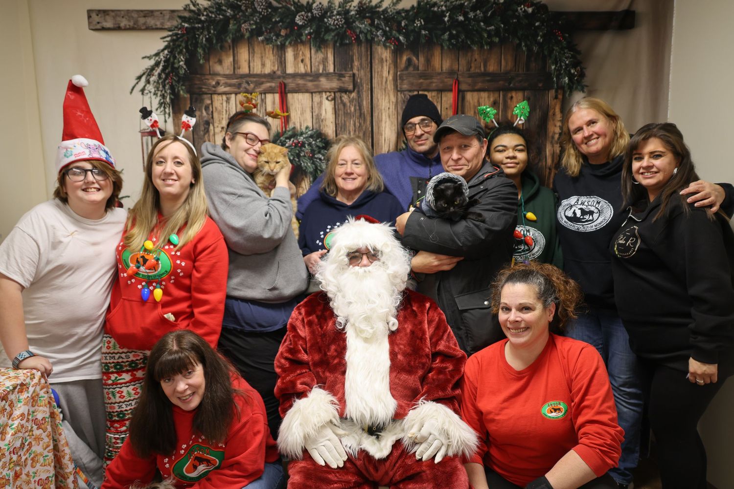 A group of people gather around Santa Claus wearing holiday attire and festive clothing for a Christmas photo.