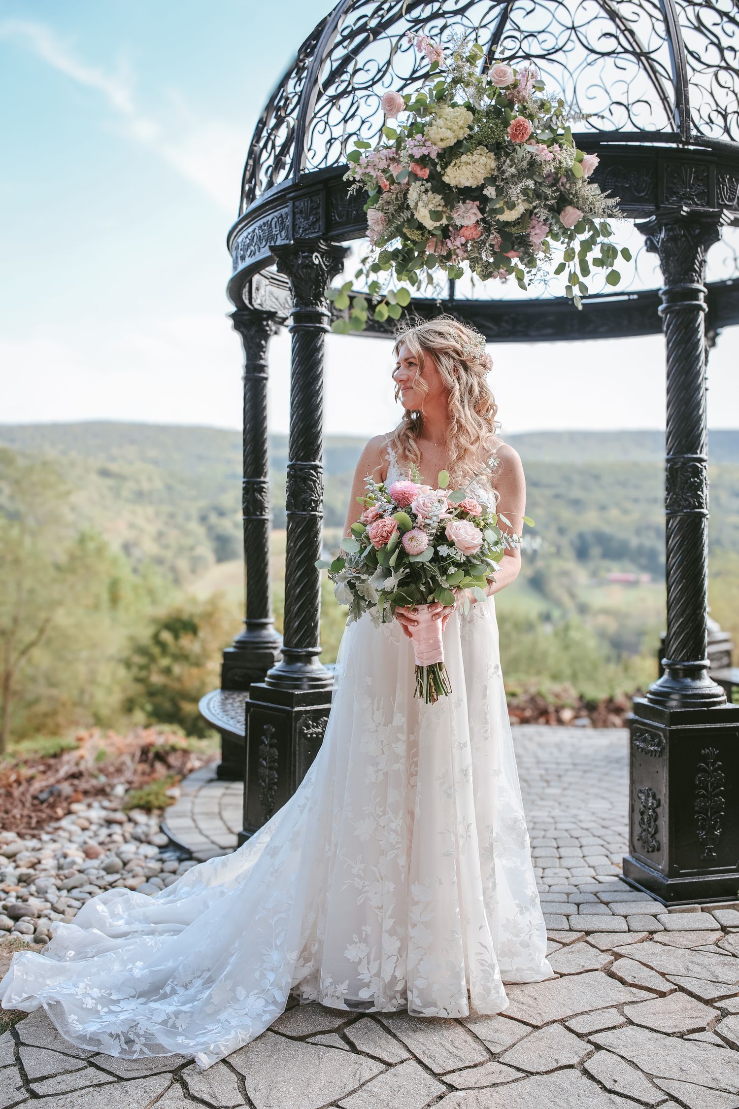 Bride in lace wedding dress poses under ornate black gazebo with floral crown and bouquet against mountain backdrop.