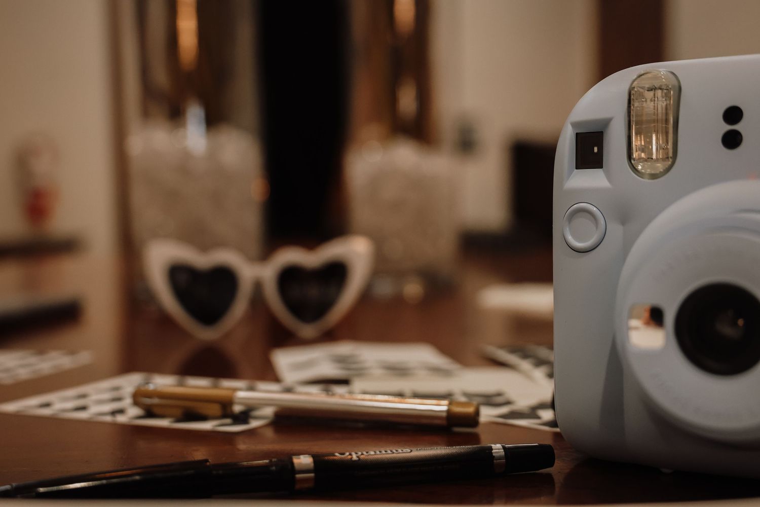 A Fujifilm Instax Mini instant camera sits on a wooden surface next to heart-shaped sunglasses and photos.