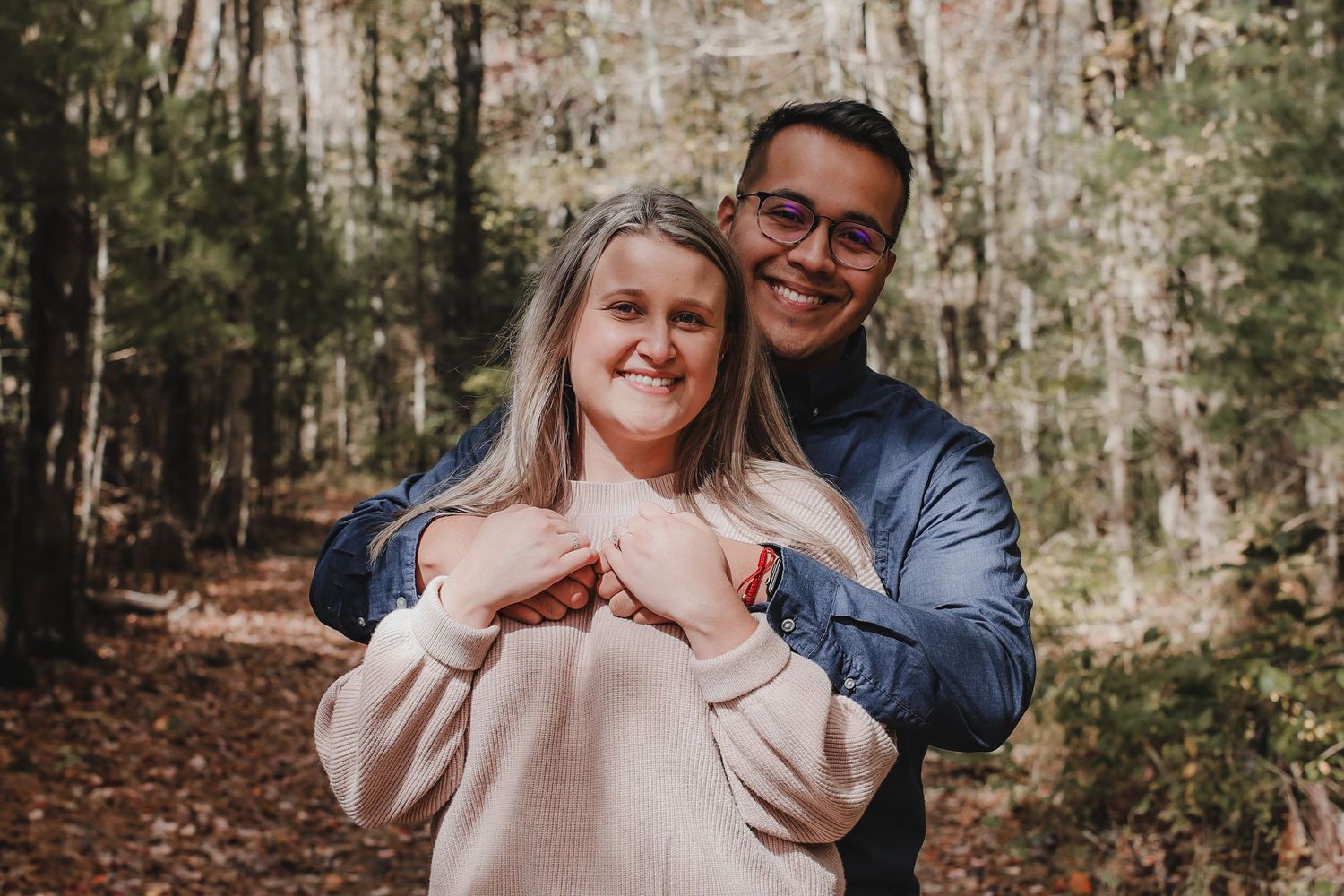 A couple embraces while standing on a forest trail surrounded by tall trees on an autumn day.