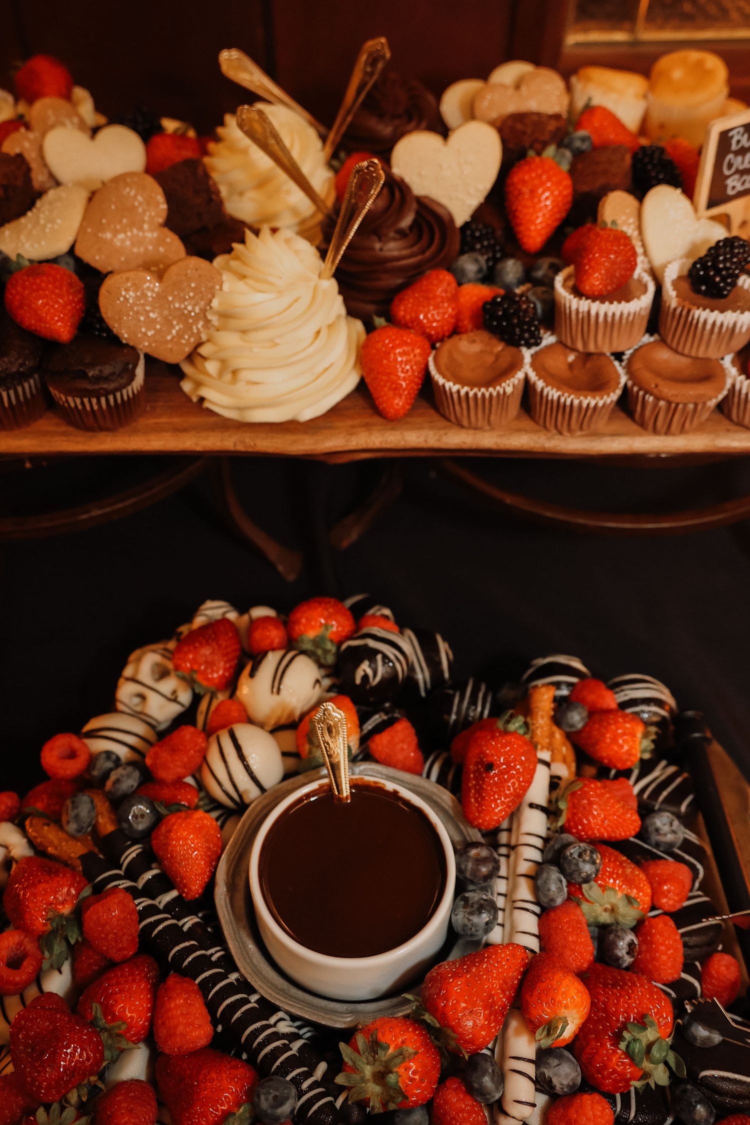 Chocolate covered strawberries and cupcakes arranged around a chocolate fondue pot on a dark surface.