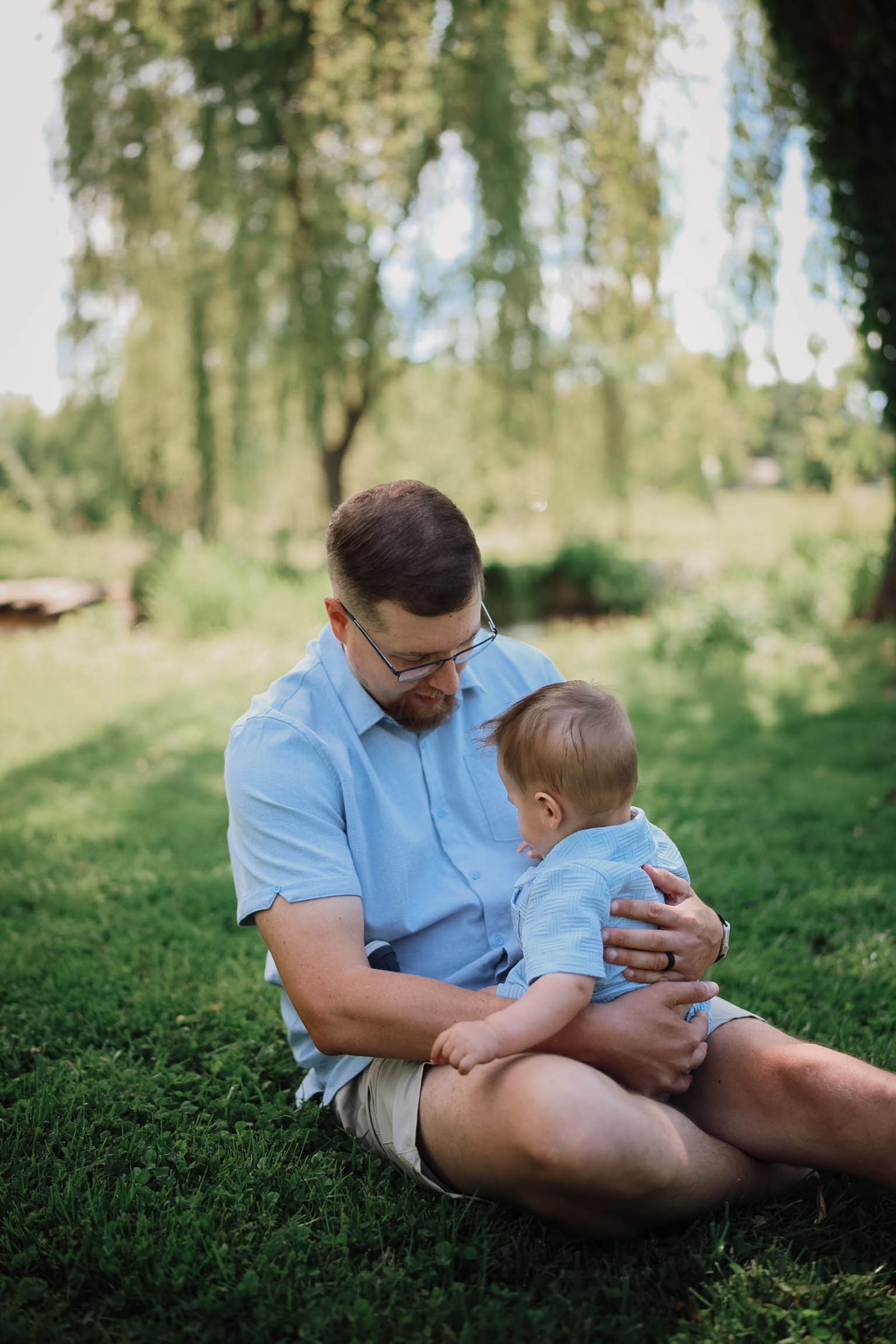 A father wearing glasses sits cross-legged on grass holding his baby in a lush green park setting.