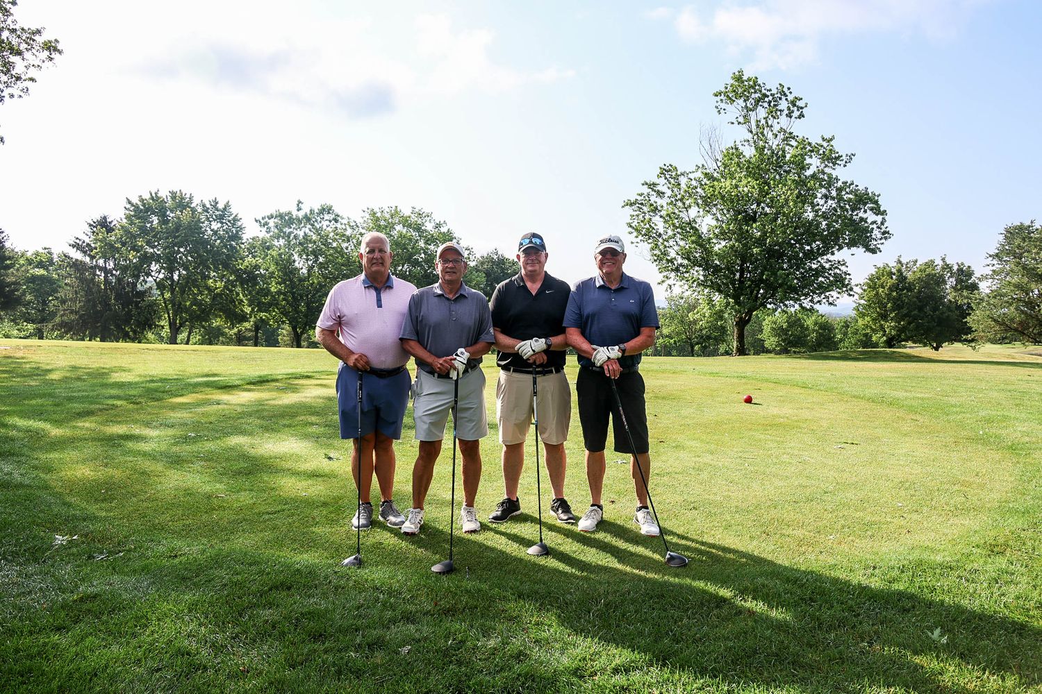Four men smiling with golf clubs on a lush green golf course on a sunny day.
