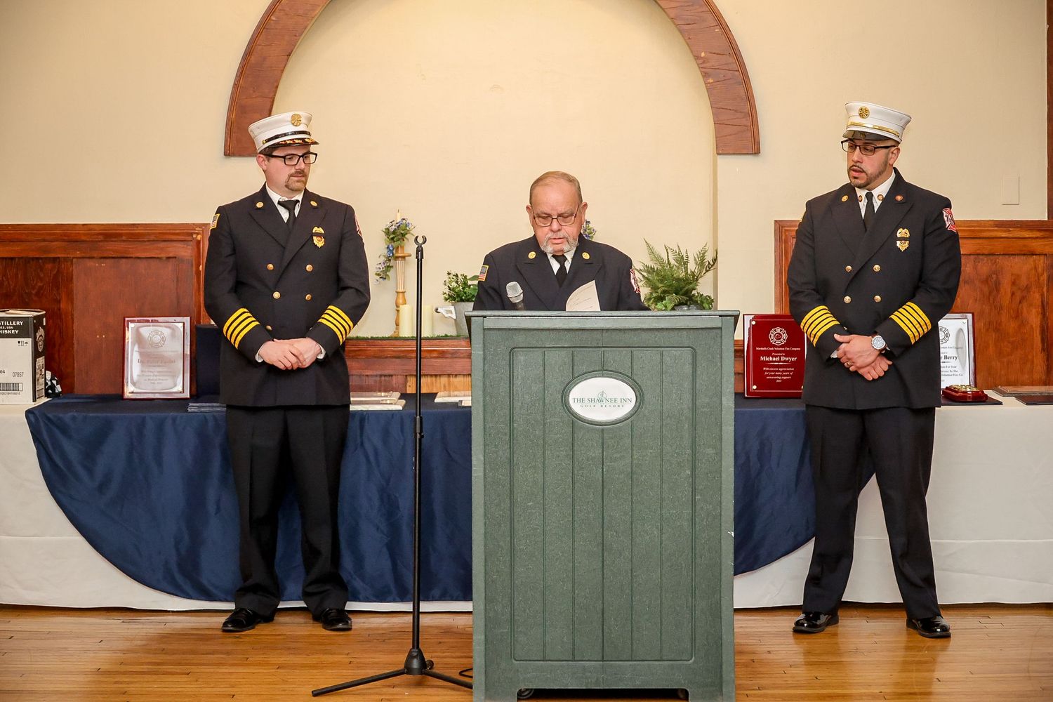 Three firefighters in formal dress uniforms stand at a podium during an official fire department awards ceremony indoors.