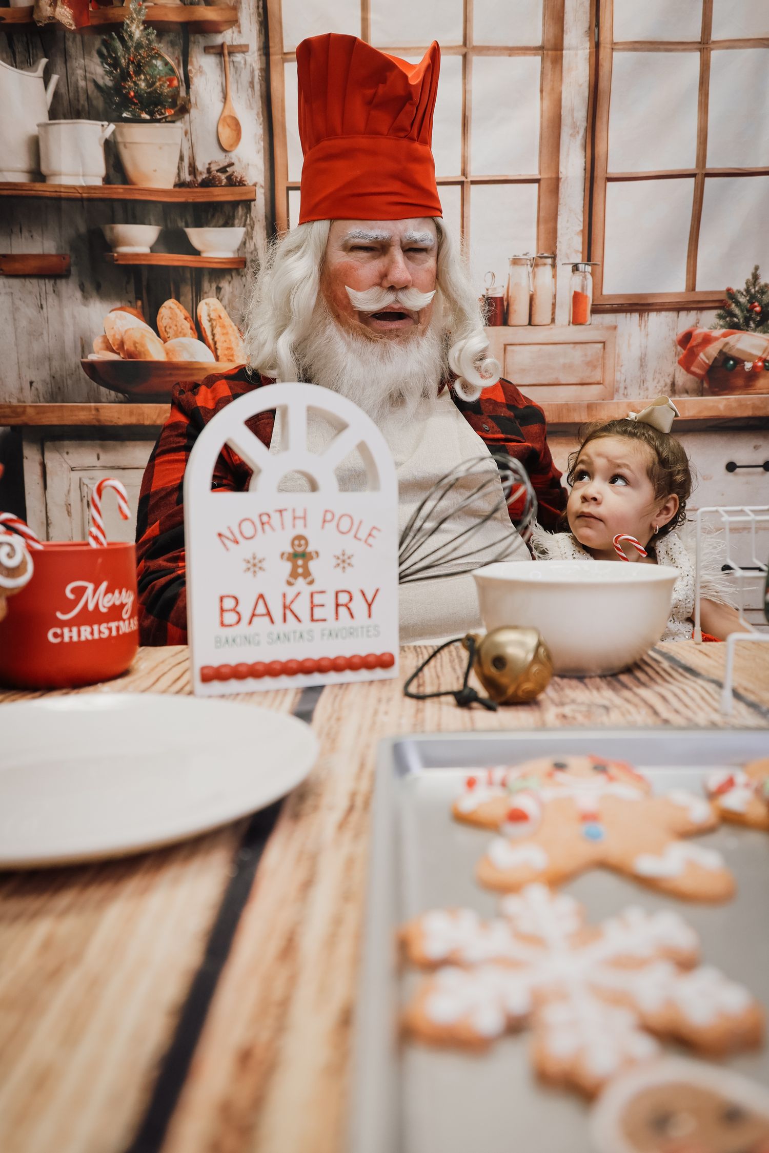 Santa Claus in red chef hat baking Christmas cookies with a young girl at North Pole Bakery kitchen.