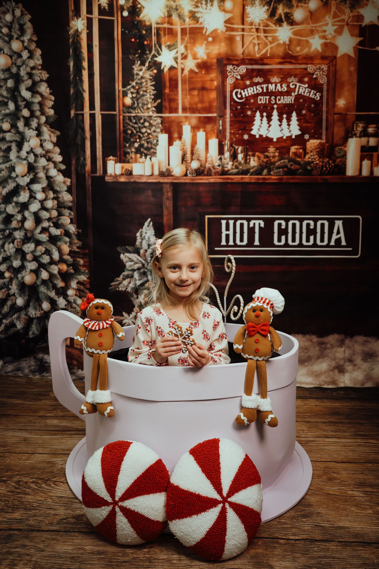 Girl sitting in giant hot cocoa mug prop with gingerbread decorations and Christmas tree backdrop.