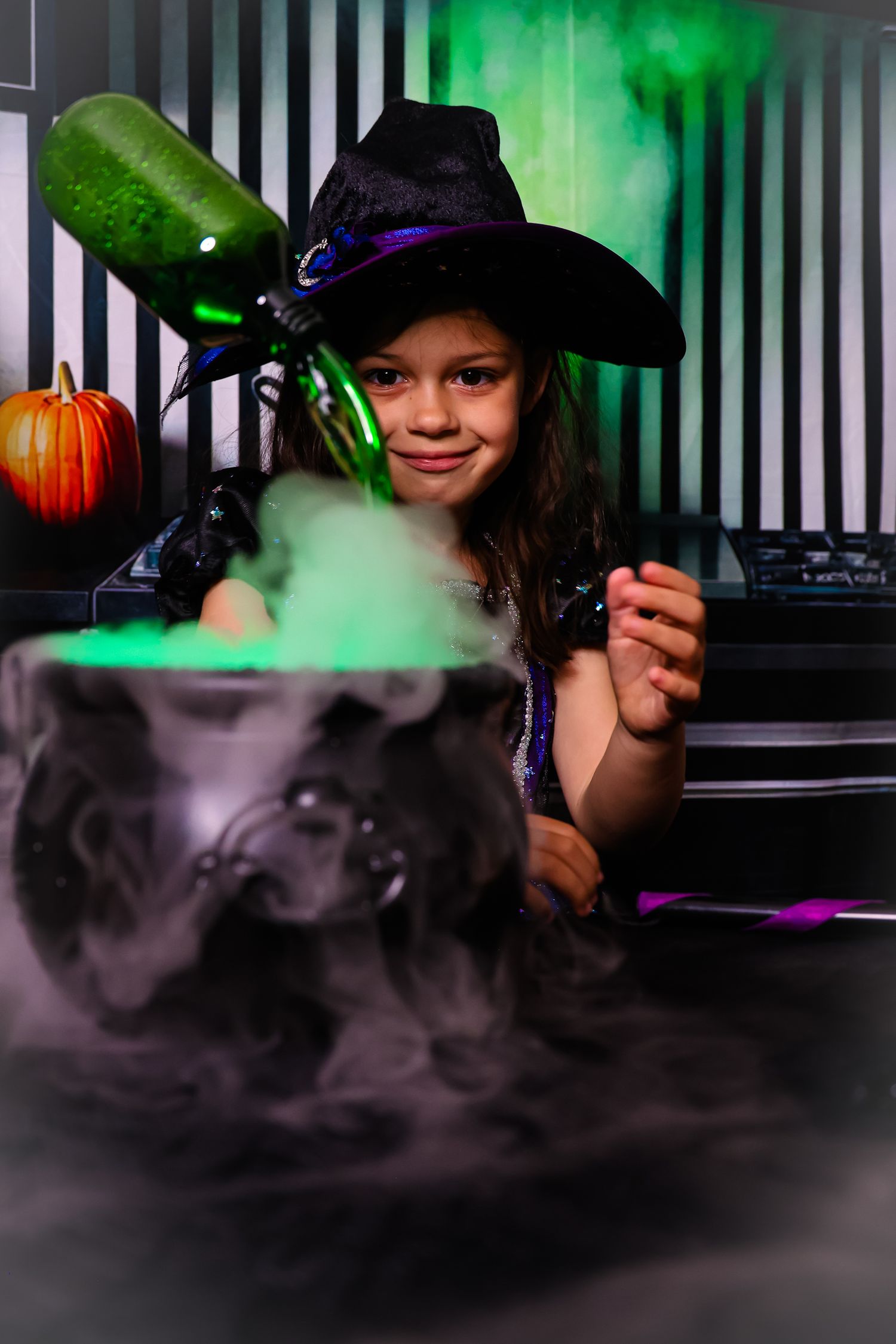 Young girl in witch hat stirs smoking Halloween cauldron with green and purple lighting effects.