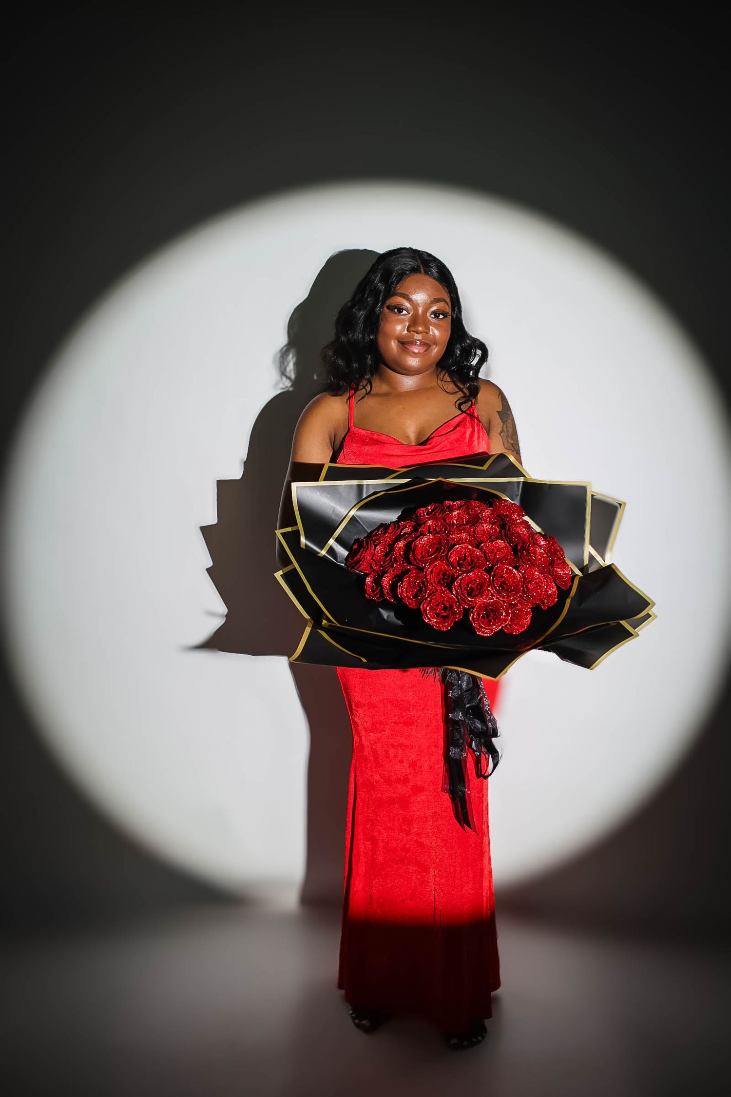 Woman in red gown holding large bouquet of red roses in black wrapping under spotlight.