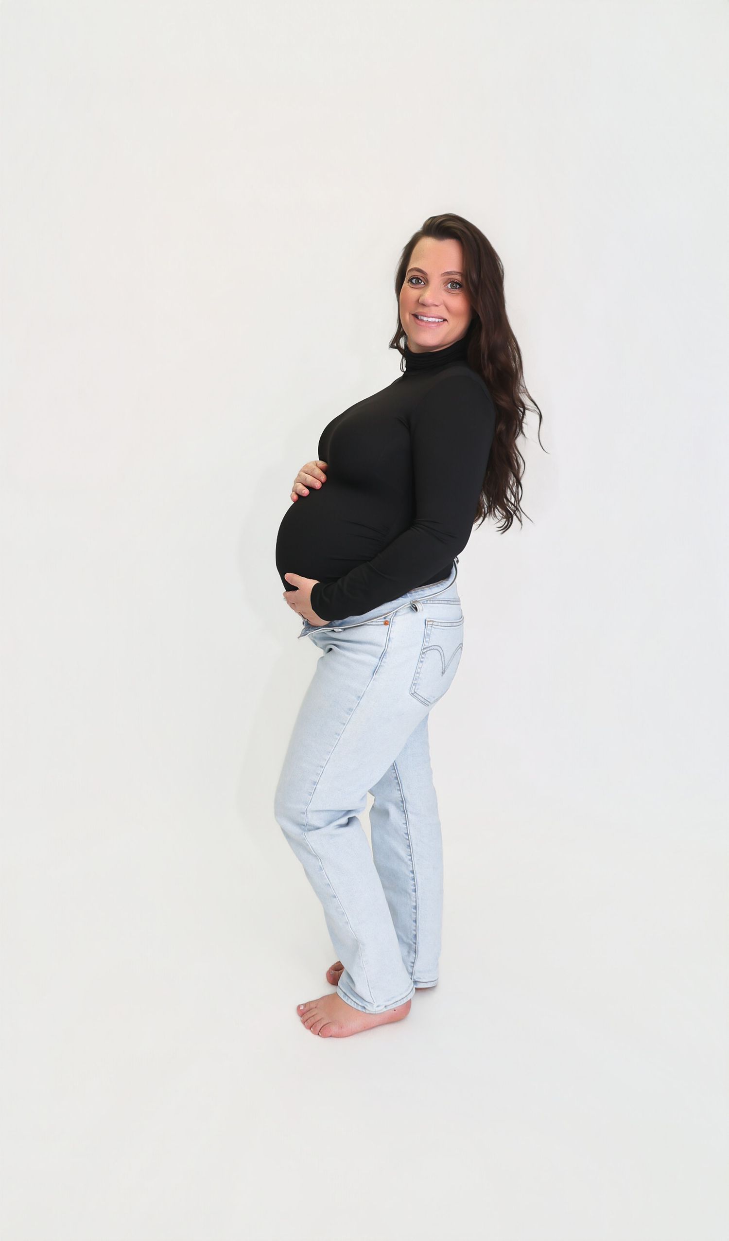 Smiling pregnant woman in black turtleneck and light blue jeans cradling baby bump, posing sideways on white background.