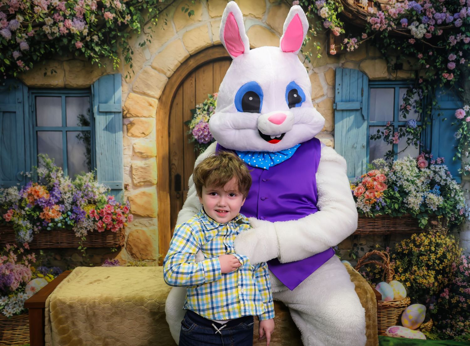 Young boy posing with Easter Bunny mascot in front of floral spring backdrop for holiday photo.